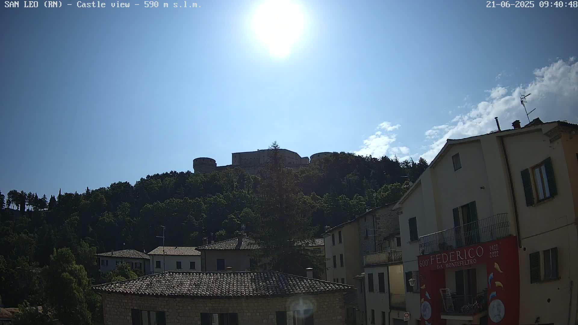 A hilltop castle is visible in the distance above a village of stone buildings under a bright, sunny sky with a few scattered clouds.