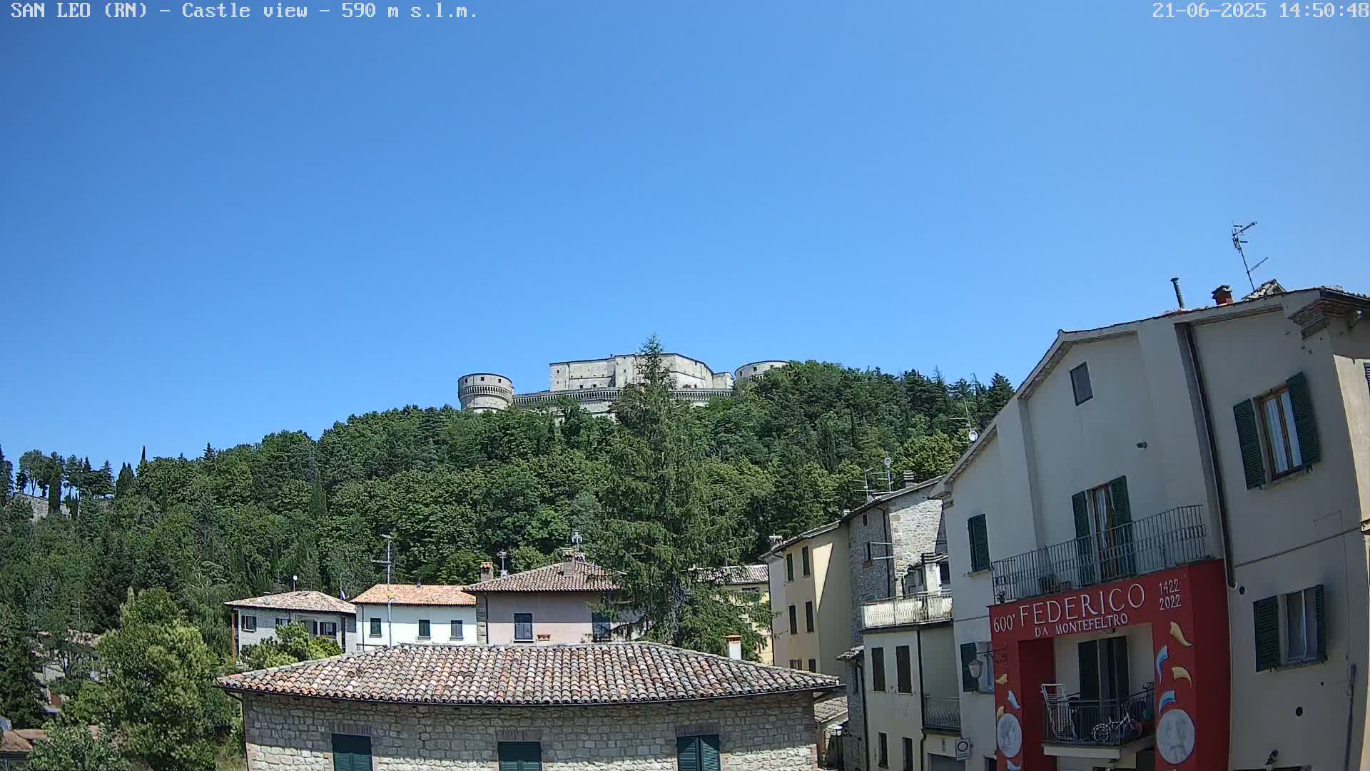 A stone castle sits atop a hill covered in lush green trees under a clear blue sky, overlooking a village of stone buildings with terracotta roofs.