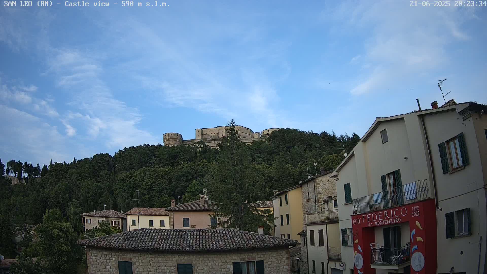 A stone castle sits atop a hill covered in lush green trees, overlooking a town of low stone buildings under a mostly clear blue sky with a few scattered clouds.