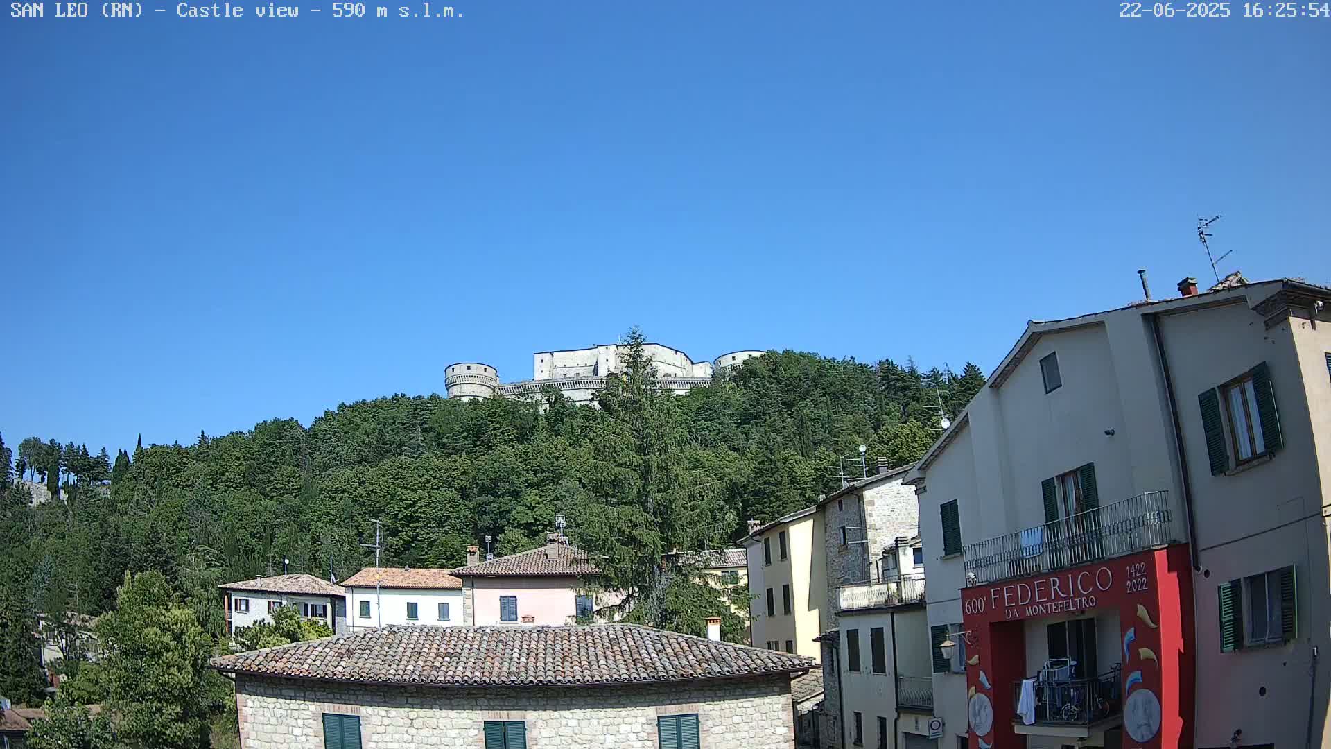 A stone castle sits atop a hill covered in lush green trees under a clear blue sky, overlooking a town with buildings of similar stone construction.