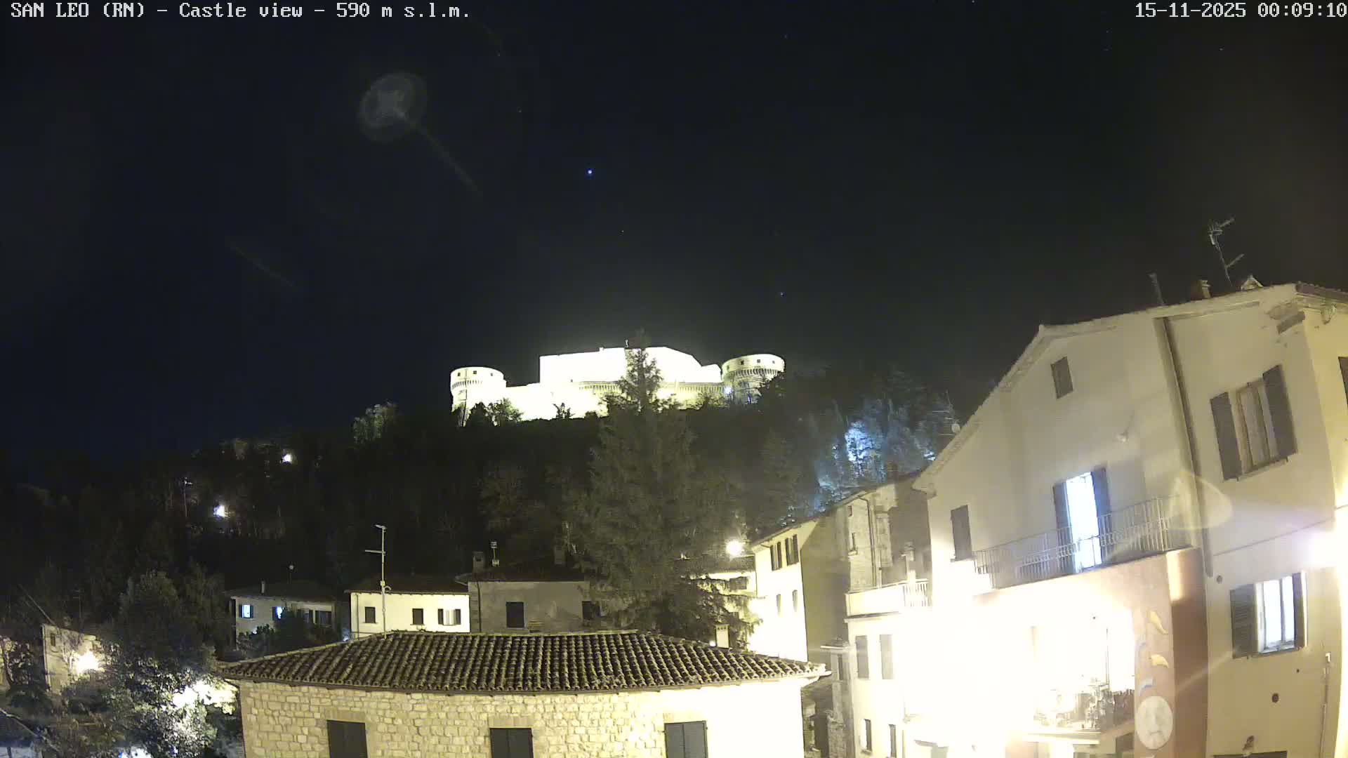 A brightly illuminated castle overlooks a town with visible residential buildings and streetlights from a dark, tree-covered hill under a clear night sky.