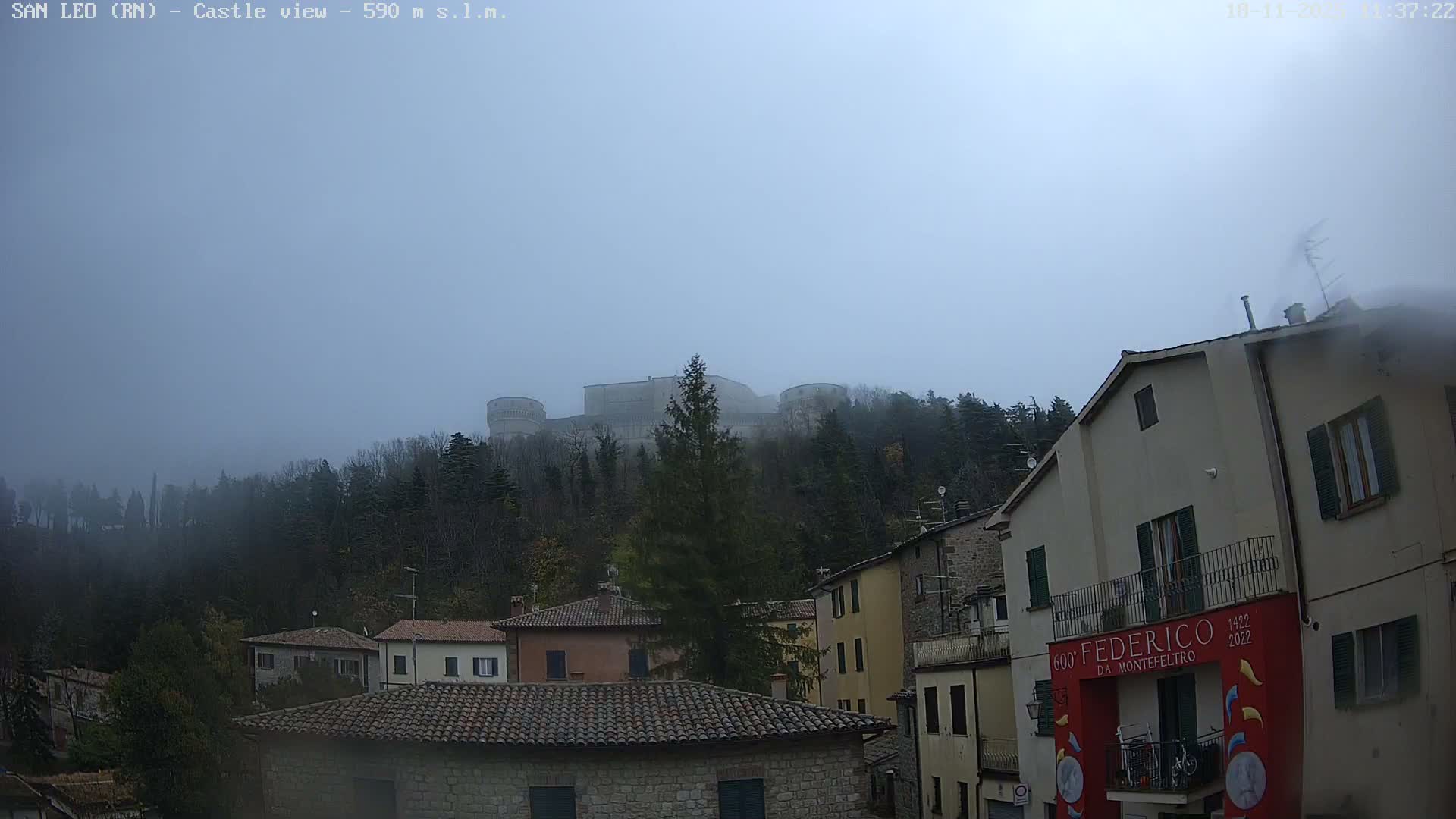 A medieval castle with round towers stands majestically atop a densely wooded hill, overlooking a quaint cluster of houses with tiled roofs under a completely overcast sky.