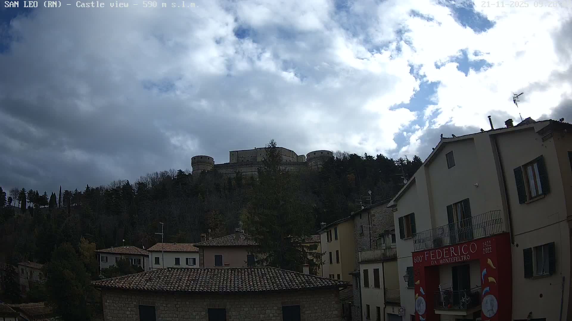 A historic fortress with multiple towers crowns a densely forested hill, overlooking a cluster of traditional buildings below, all under a partly cloudy sky with intermittent sunlight.