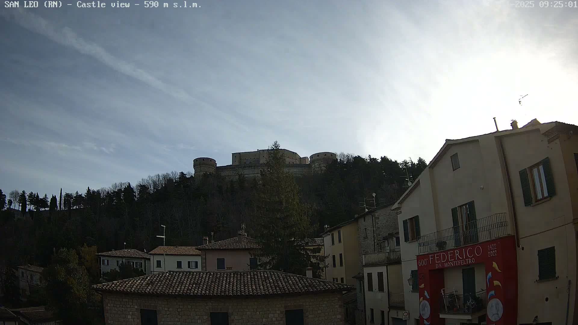 A historic fortress crowns a tree-covered hill overlooking a cluster of houses in a town below, all illuminated by bright sunshine on a partly cloudy day.