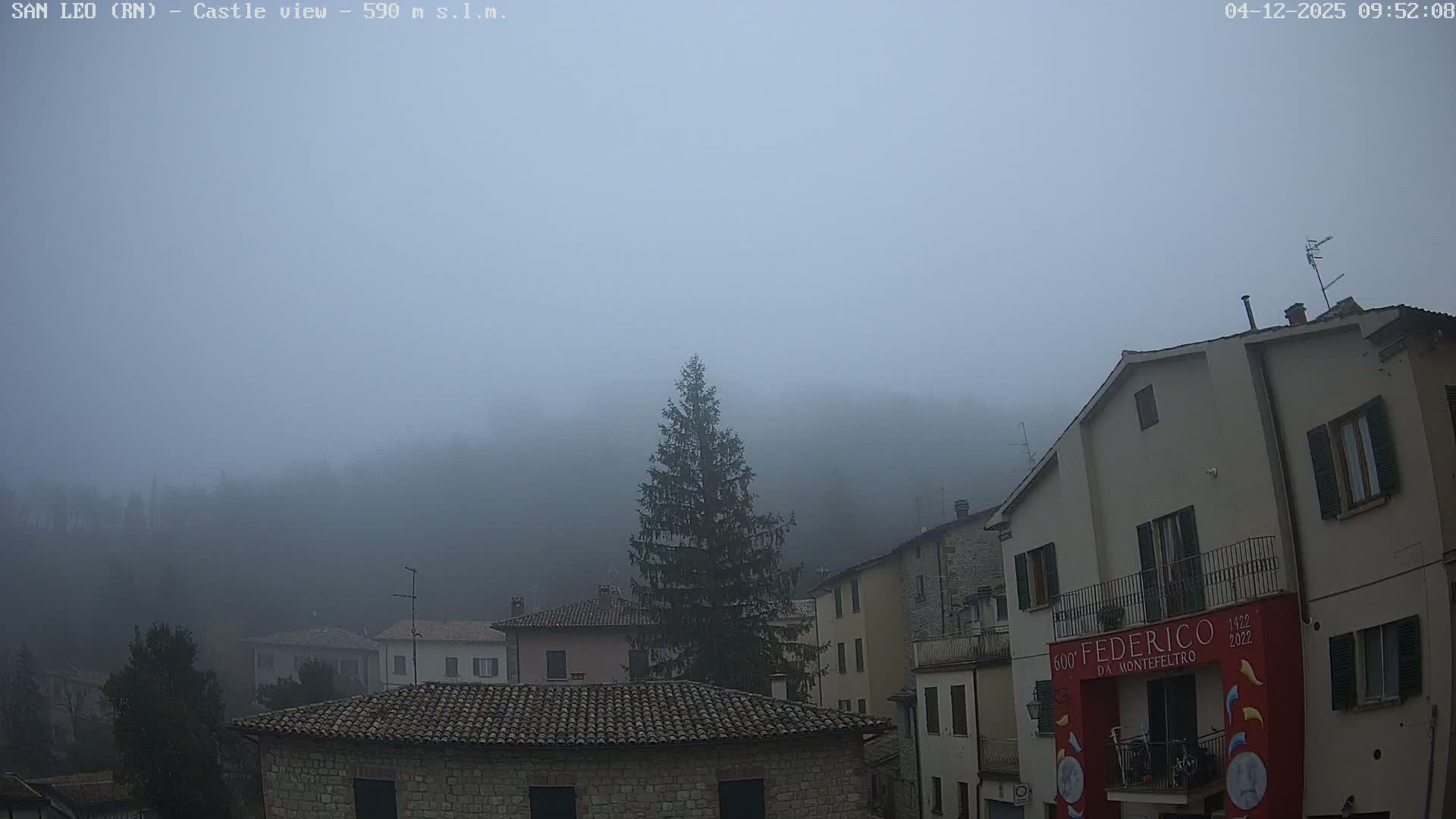 Dense fog blankets a town of stone and stucco buildings with tiled roofs and a prominent evergreen tree, as distant hillsides fade into the heavy mist.