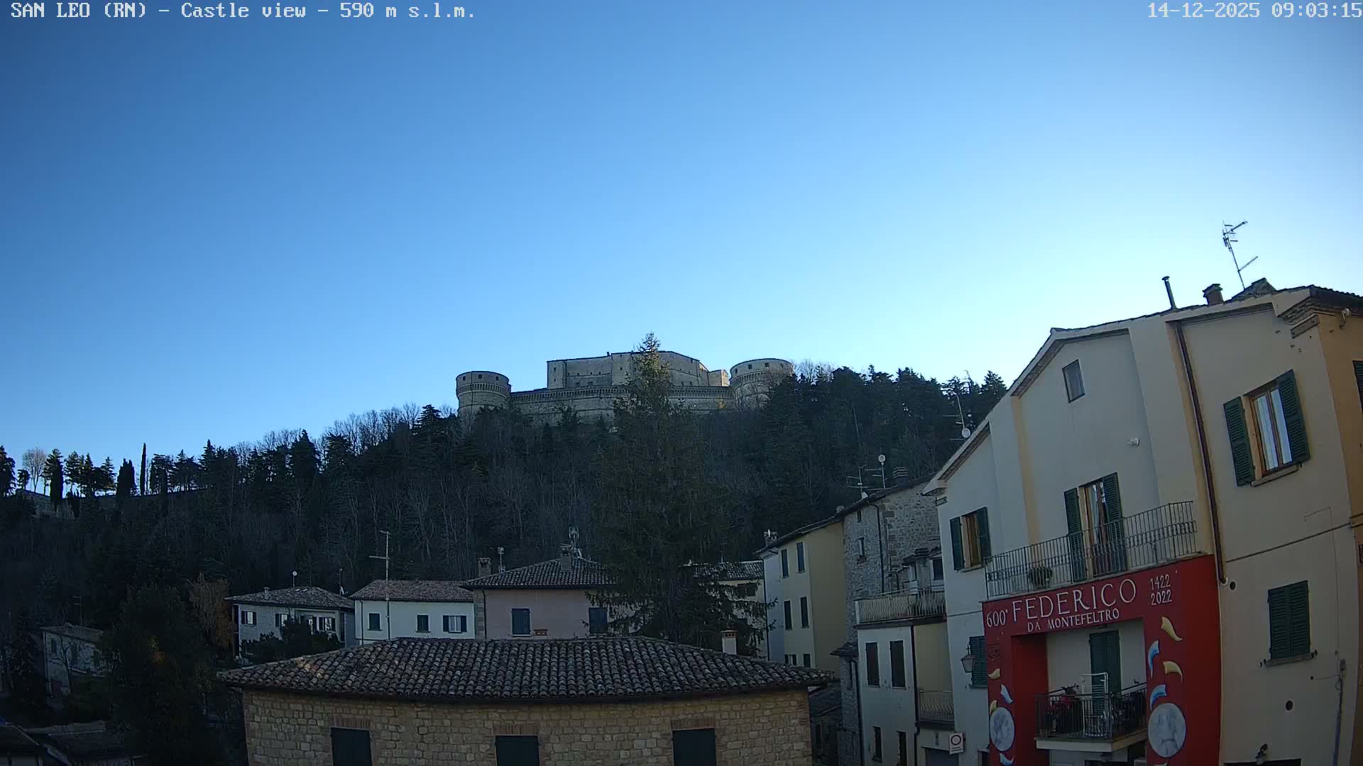 Under a clear blue sky, a large stone castle crowns a densely wooded hill, overlooking a cluster of residential buildings with tiled roofs in the foreground.