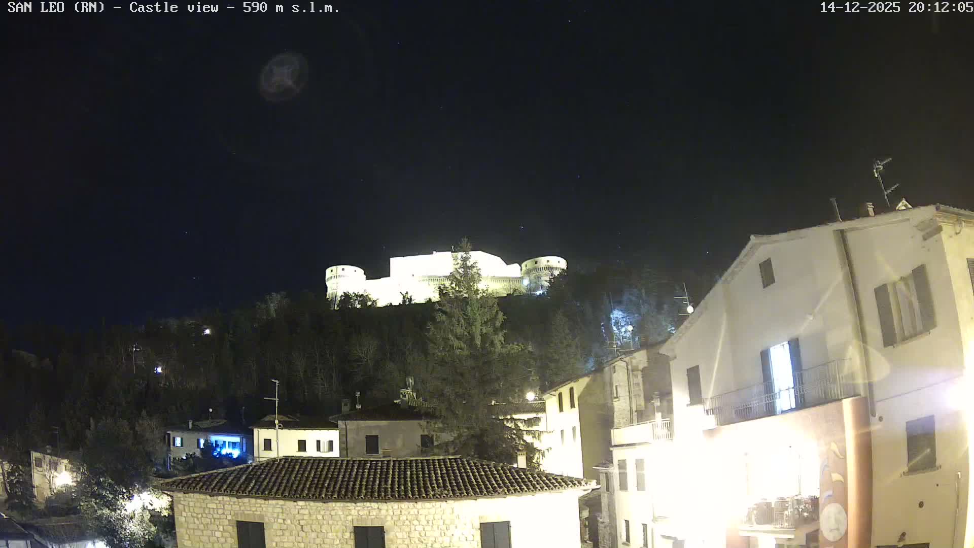 Under a clear blue sky, a large stone castle crowns a densely wooded hill, overlooking a cluster of residential buildings with tiled roofs in the foreground.