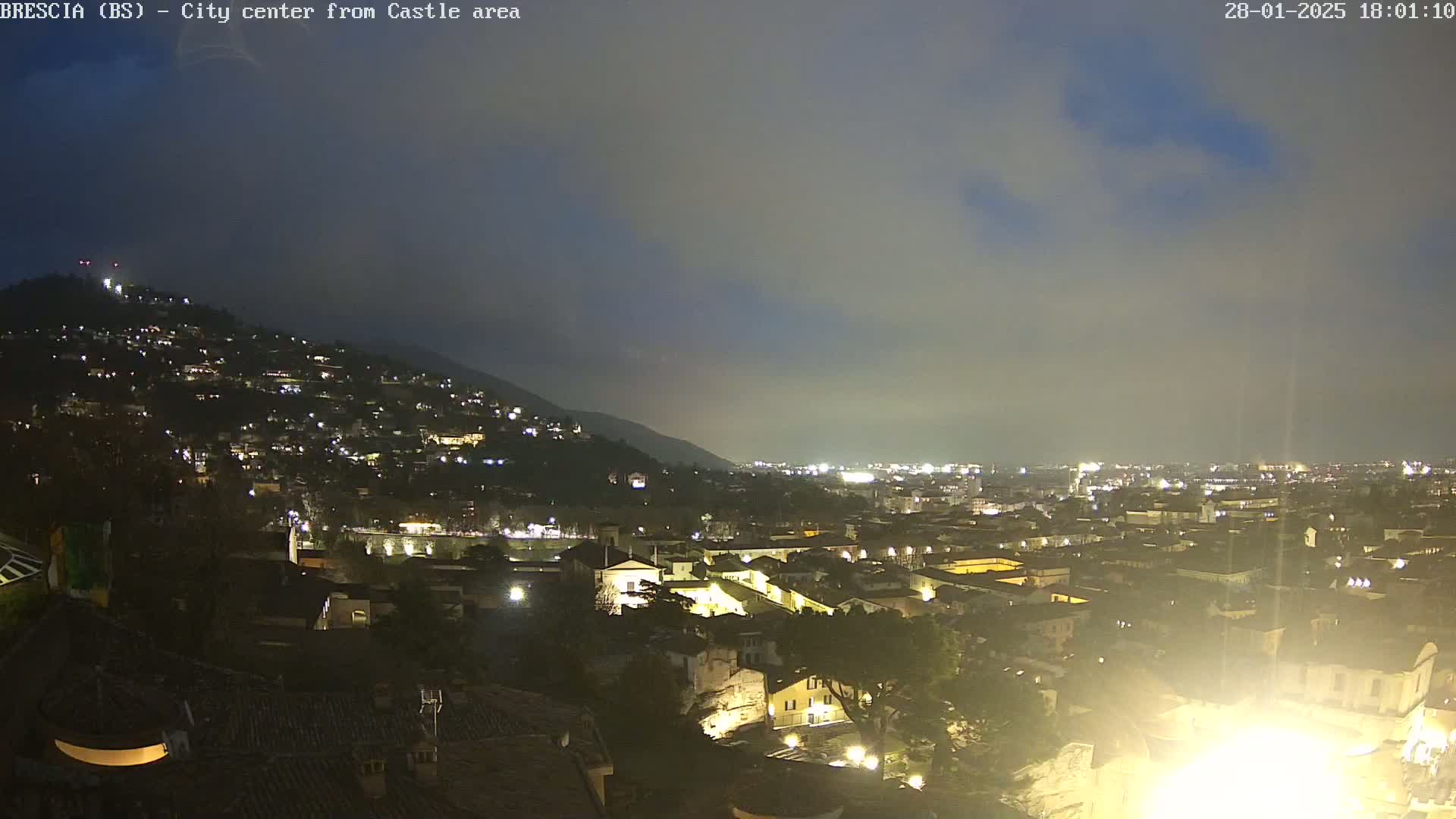 A nighttime view of a city nestled in a valley under a cloudy sky, illuminated by numerous lights.