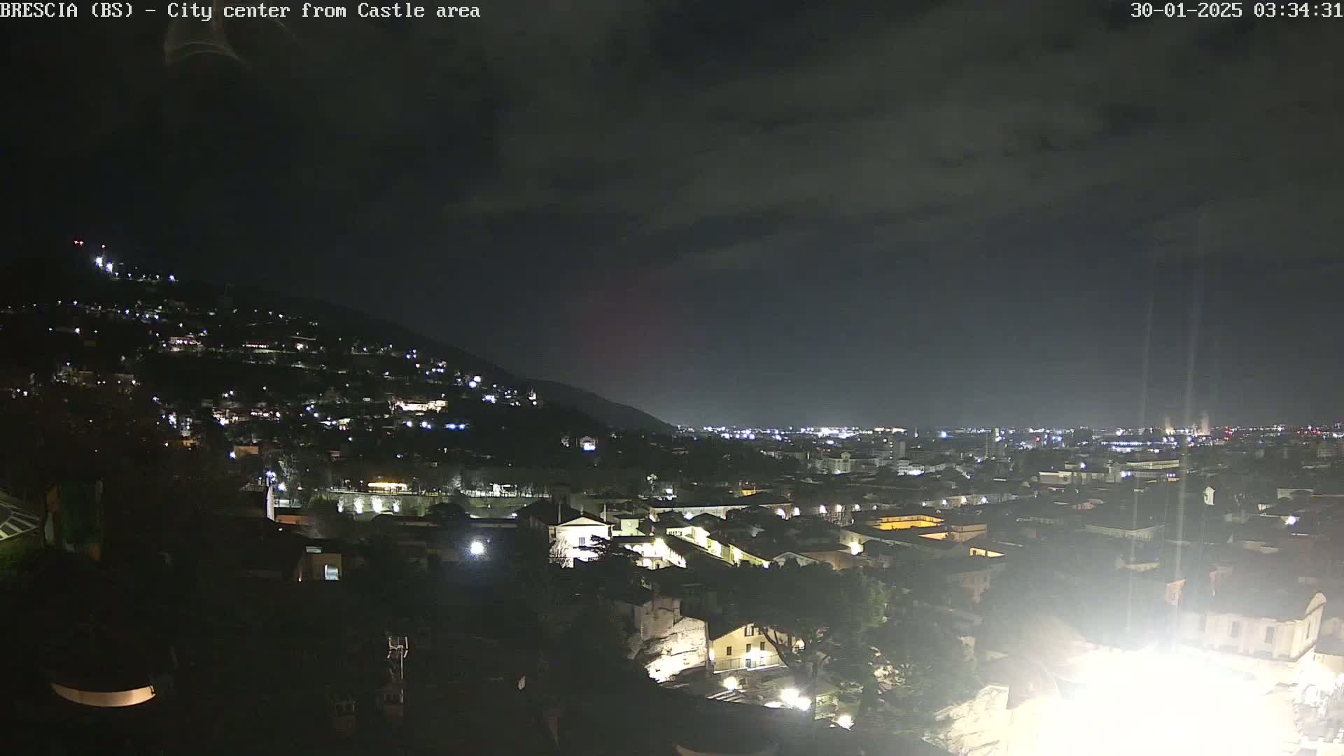 A nighttime view of a city nestled in a valley under a mostly cloudy sky, with numerous lights illuminating buildings and streets.