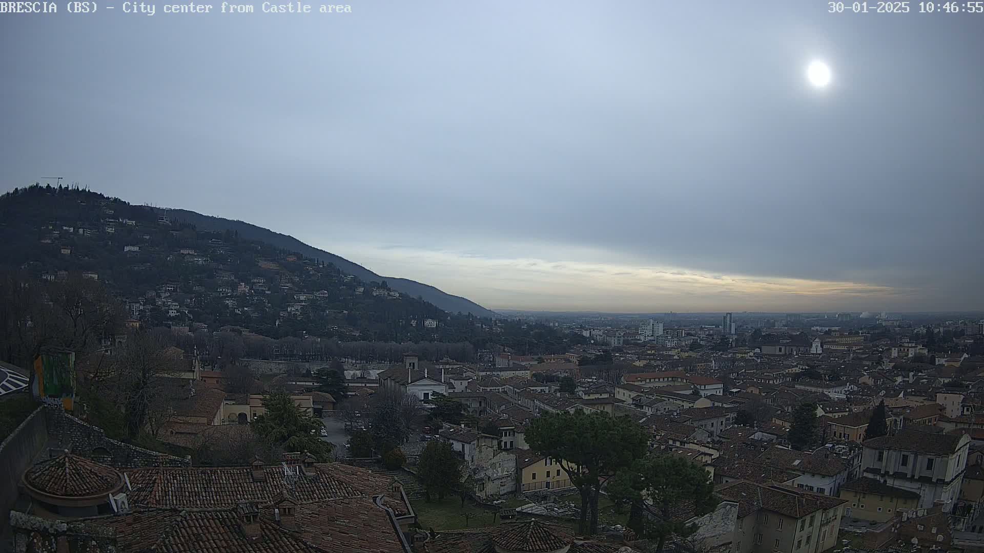 A sunlit cityscape stretches out below a mostly cloudy sky, nestled between a hill covered in buildings and vegetation and a broader, flatter expanse of the city in the distance.