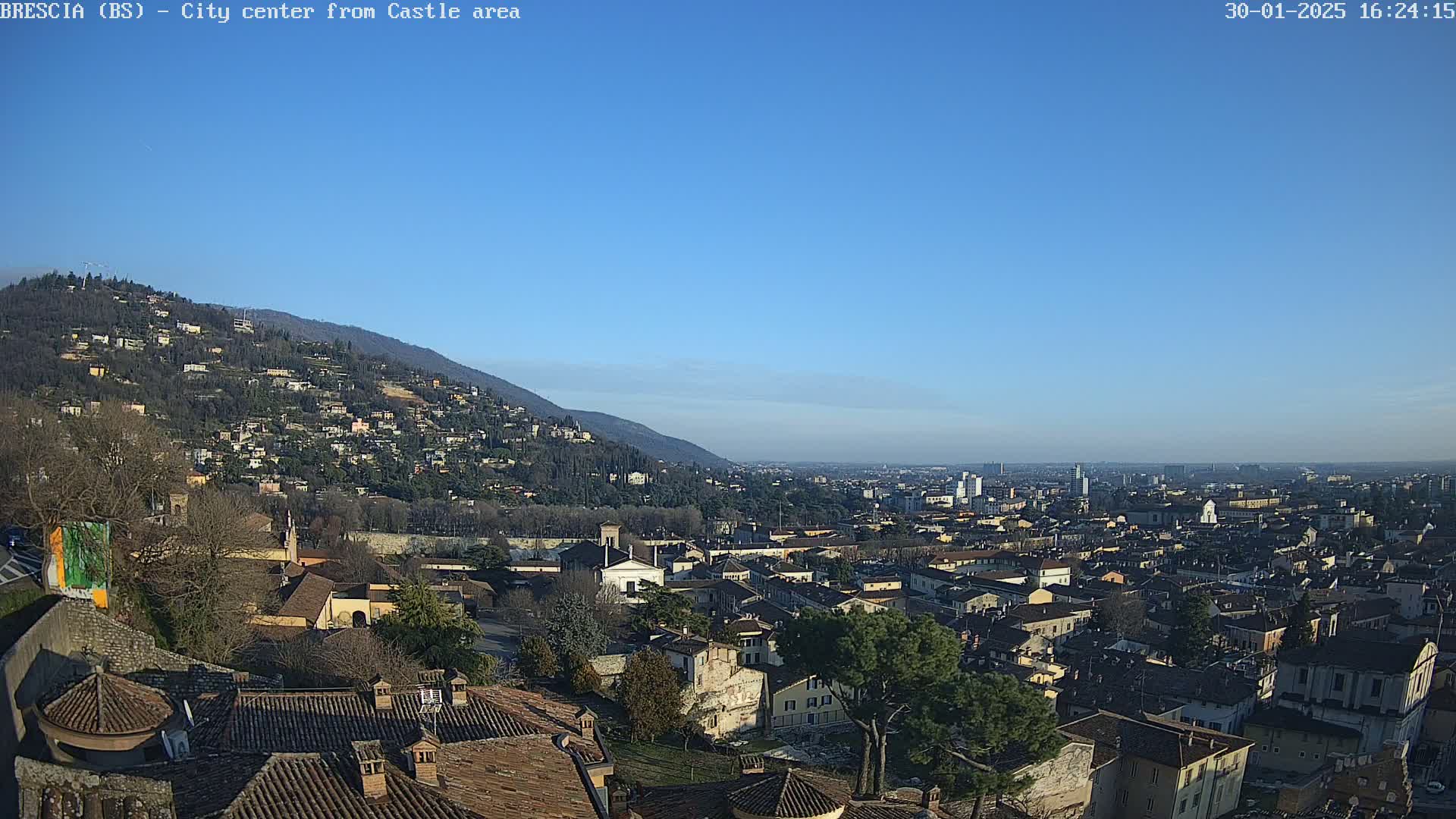 A panoramic view of a city nestled in a valley, under a clear blue sky, shows a hillside dotted with buildings in the background.