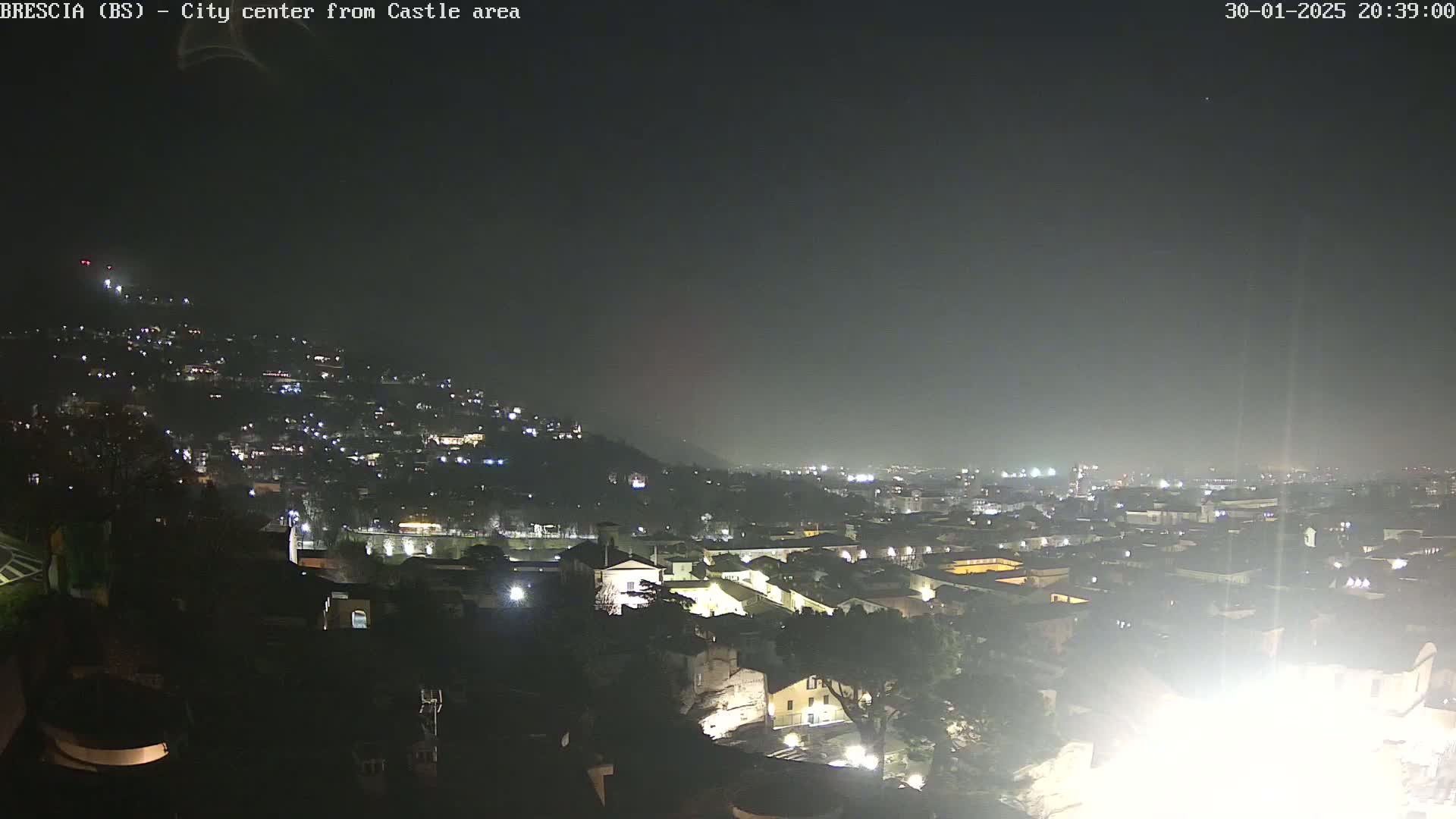 A nighttime, hazy view of a city illuminated by lights, seen from a hilltop vantage point.