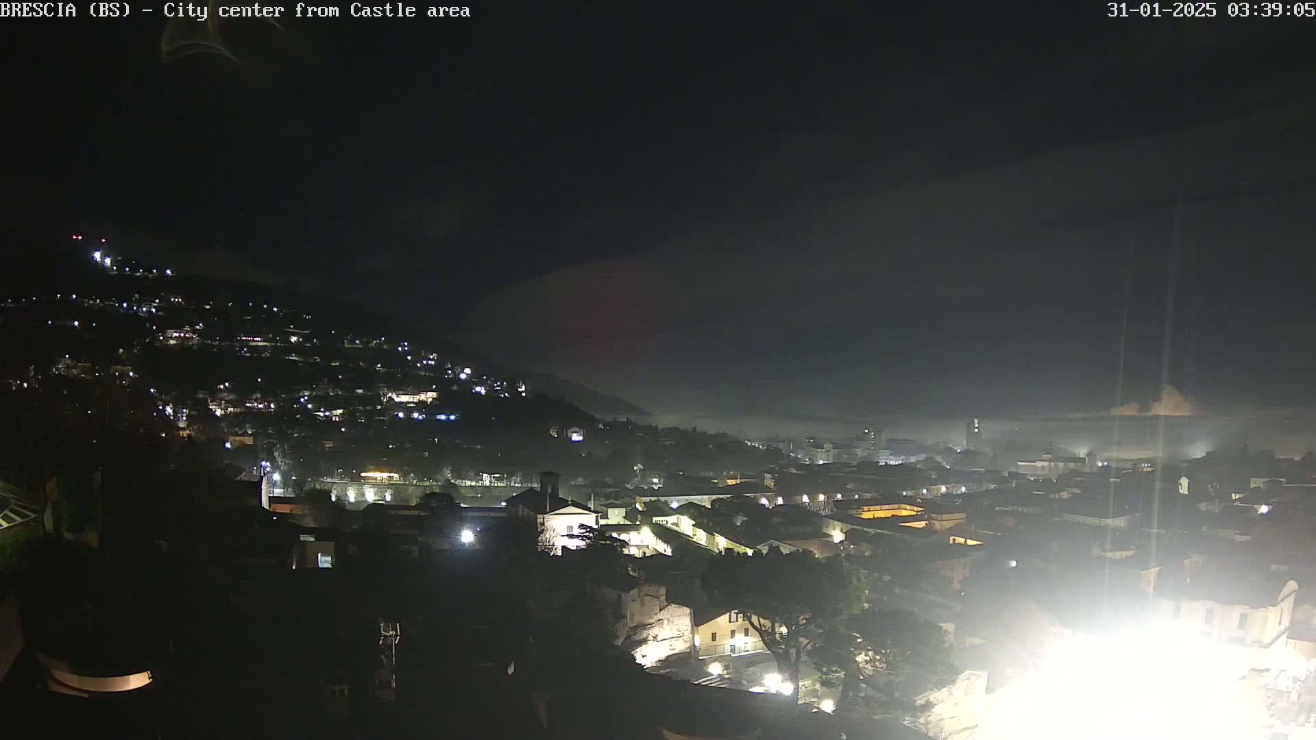 A nighttime, foggy view of a city nestled on a hillside, with scattered lights illuminating the buildings and streets.
