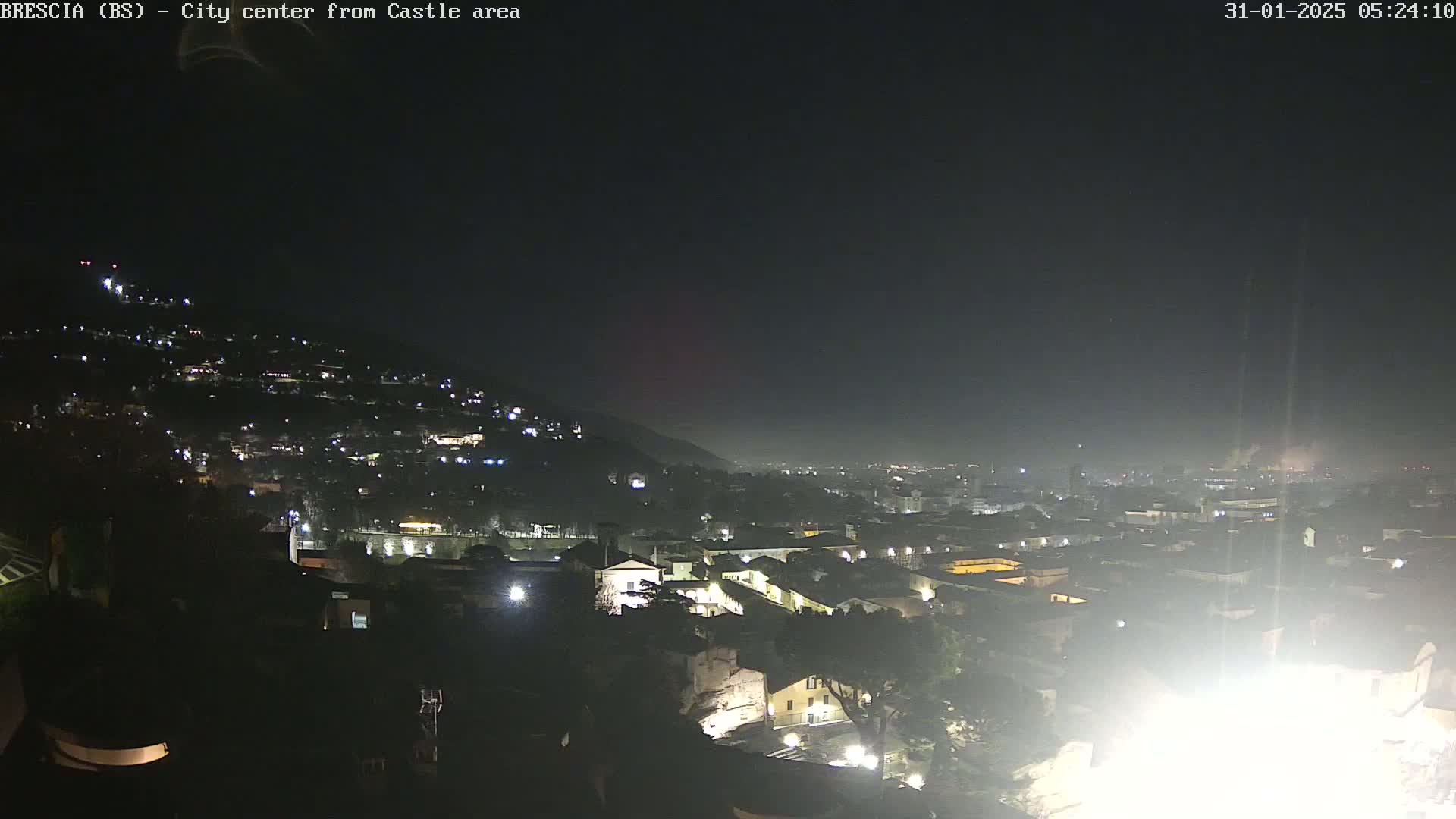 A nighttime view of a city nestled on a hill, illuminated by scattered lights under a dark, hazy sky.