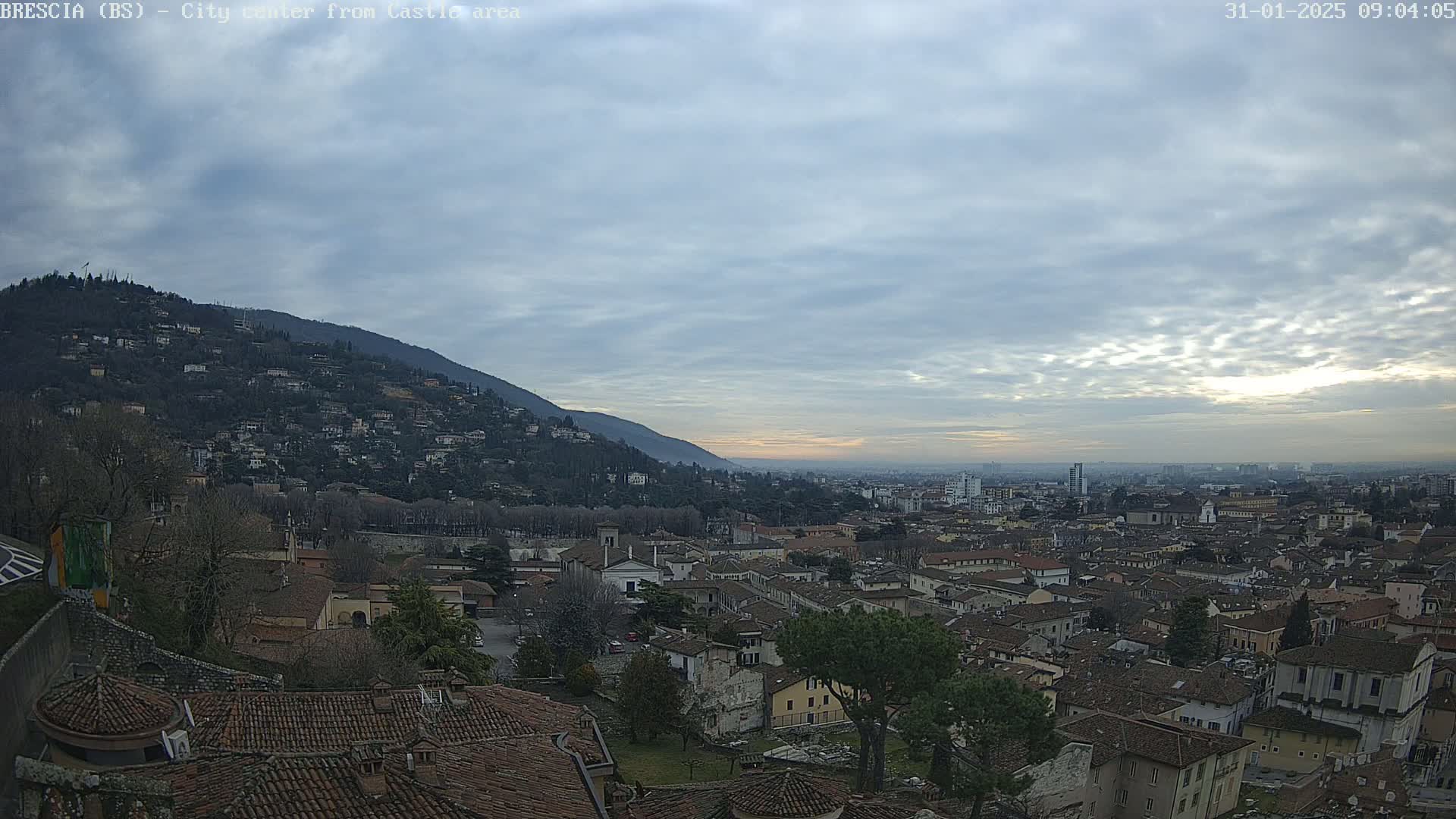 A sprawling city landscape nestled against a hill, under a mostly cloudy sky with hints of sunrise or sunset.