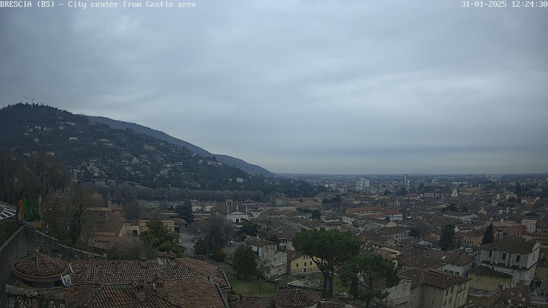 A sprawling city with red-tiled roofs nestled at the base of a hill, under a cloudy sky.