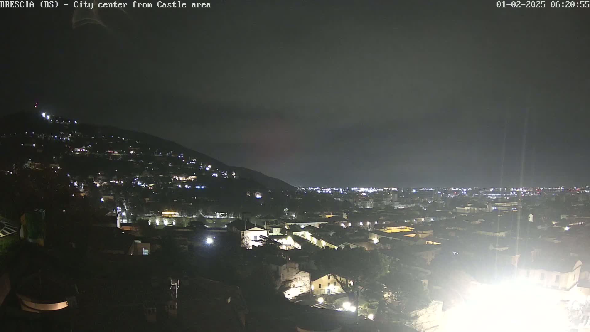 A nighttime view of a city illuminated by lights, nestled against a dark hill under a cloudy sky.