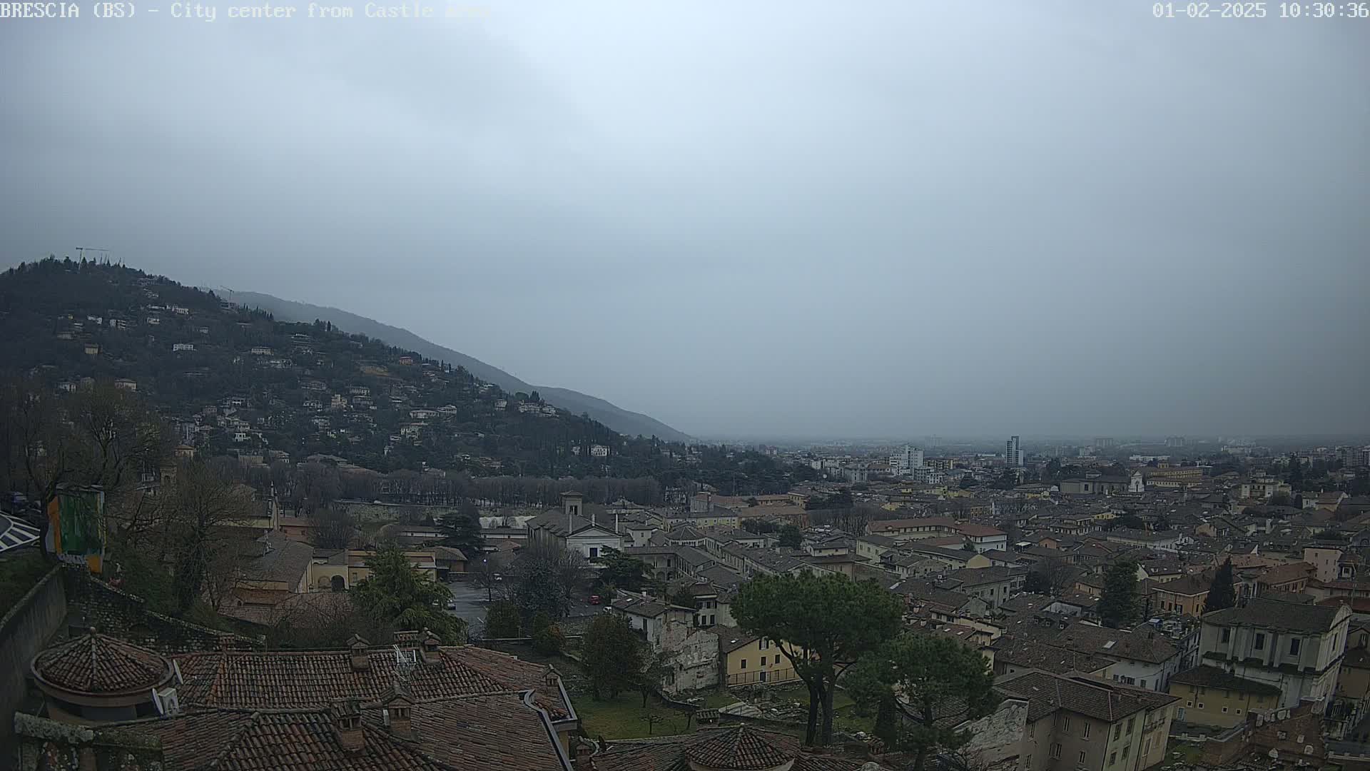 A hazy, overcast view of a city nestled in a valley, with a hill covered in buildings in the foreground and a larger city sprawling in the distance.