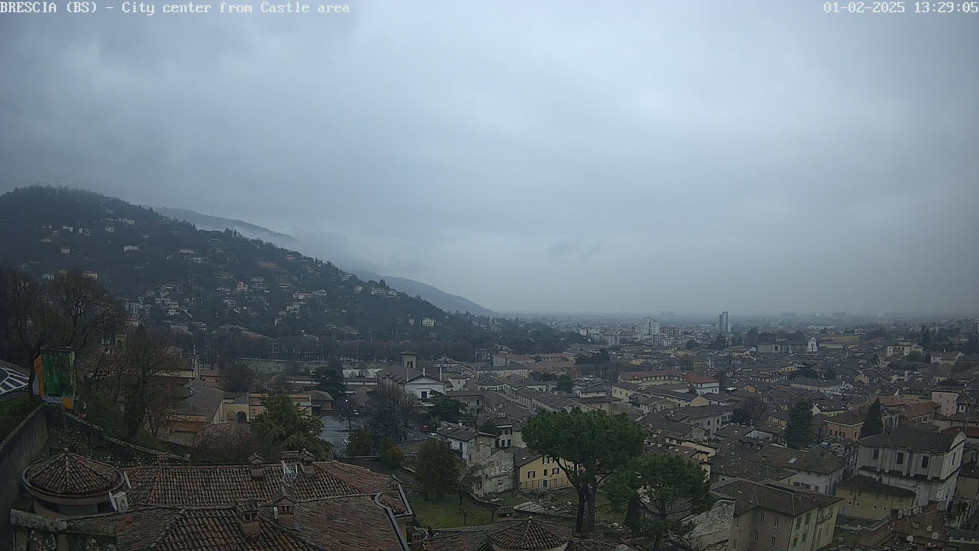 A hazy, overcast view shows a city nestled in a valley between hills, with many buildings with reddish-brown roofs visible.