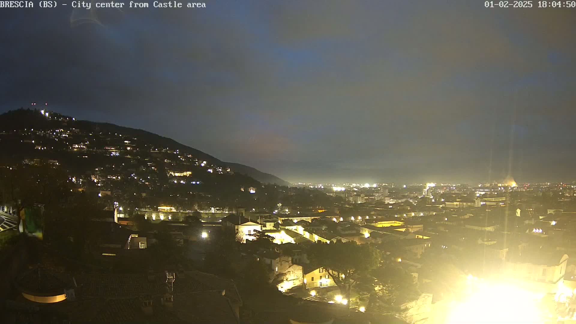 A nighttime view of a city illuminated by lights, nestled against a hill under a cloudy sky.