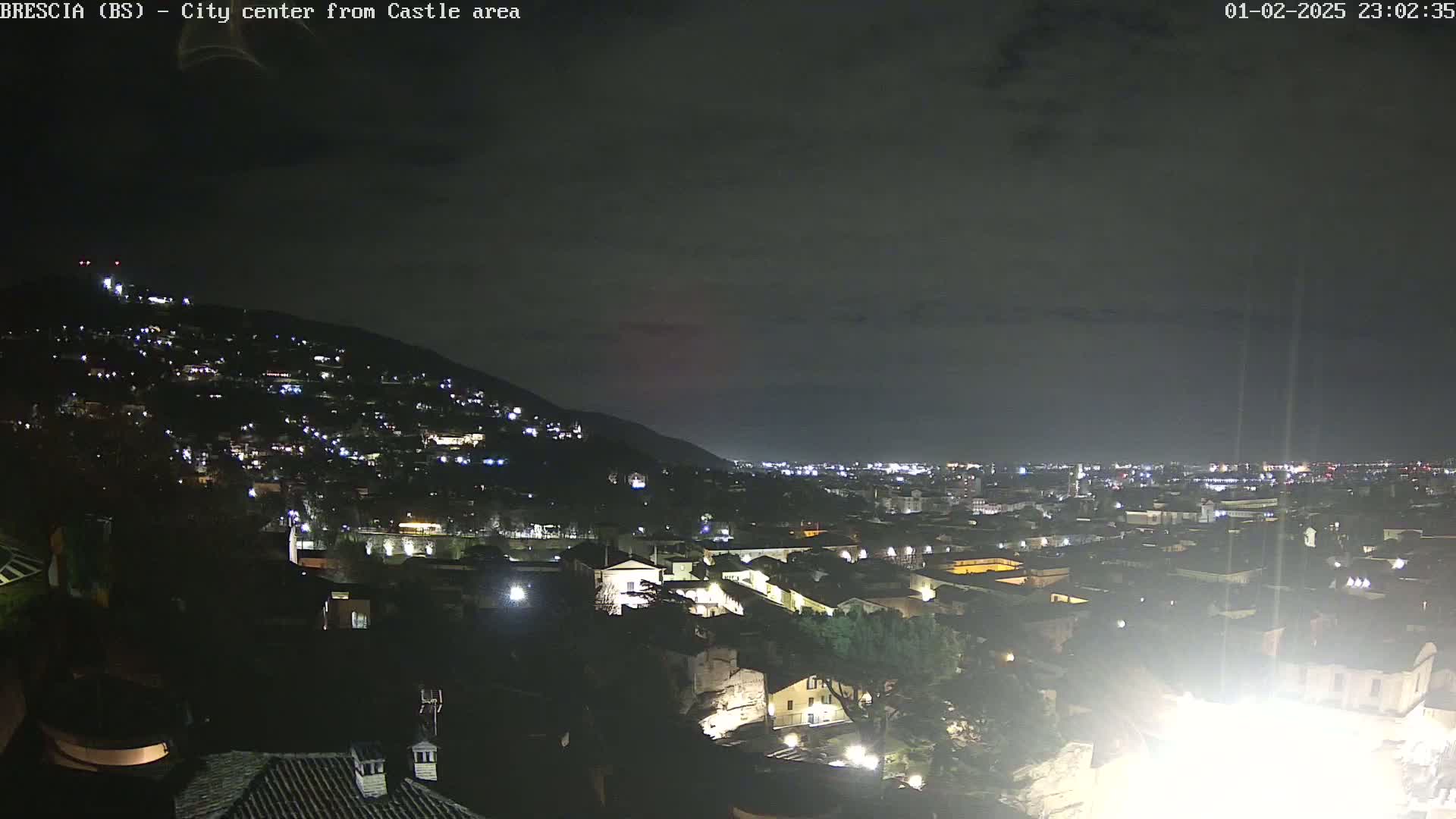 A nighttime view of a city nestled against a hill, illuminated by numerous lights under a dark, cloudy sky.