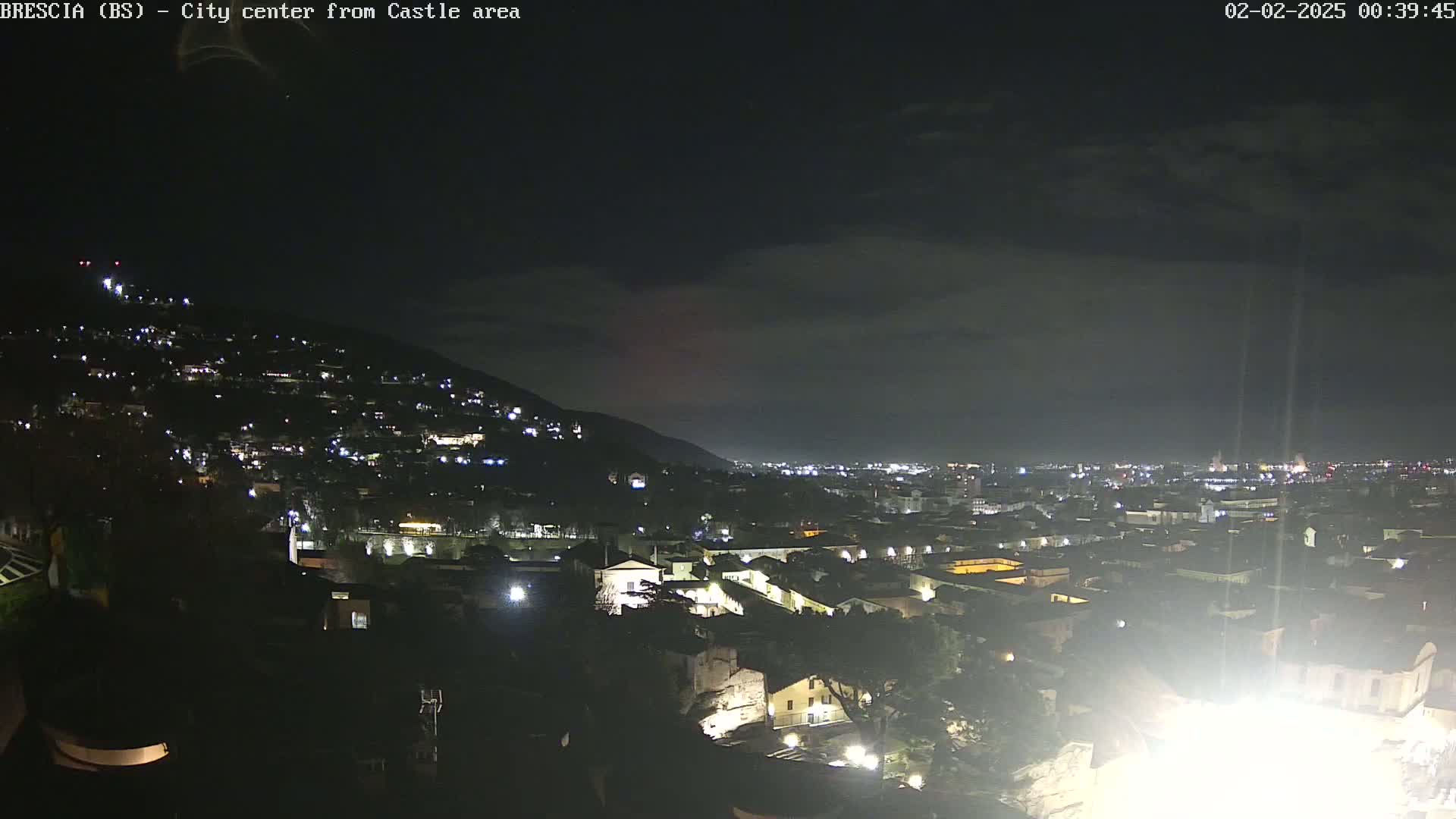 A nighttime view of a city nestled against a hill, under a mostly cloudy sky.