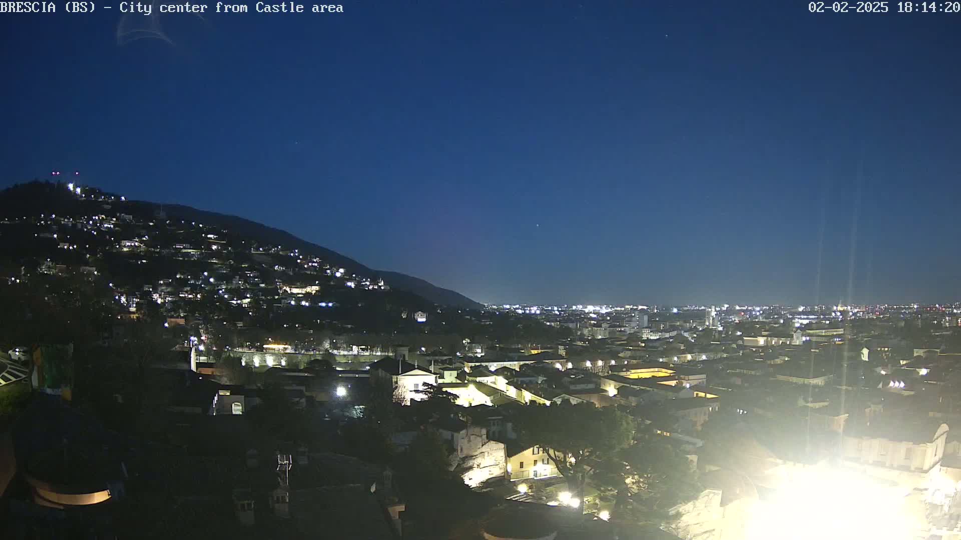A nighttime view of a city illuminated by lights, nestled against a dark hill under a clear, dark blue sky.