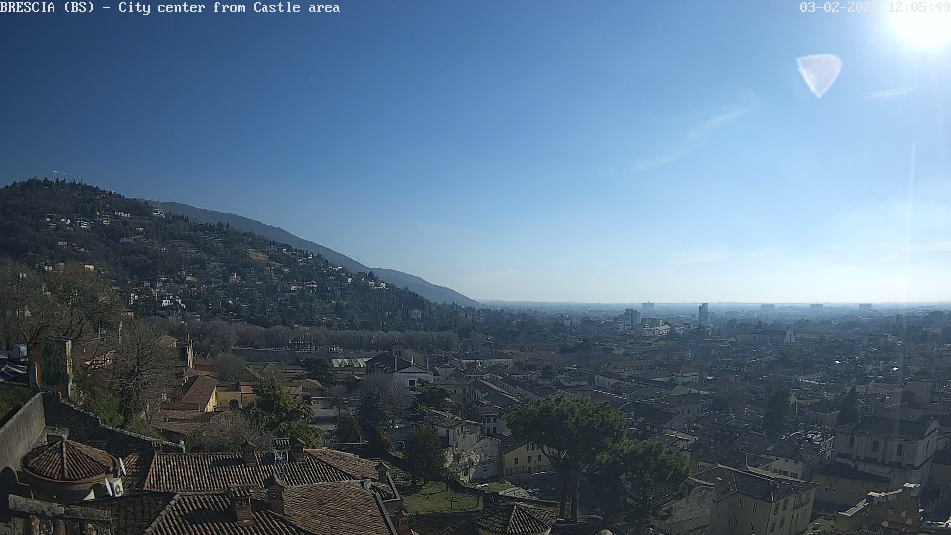 A sunny day reveals a sprawling cityscape nestled beside a hill, with a distant mountain range visible on the horizon.