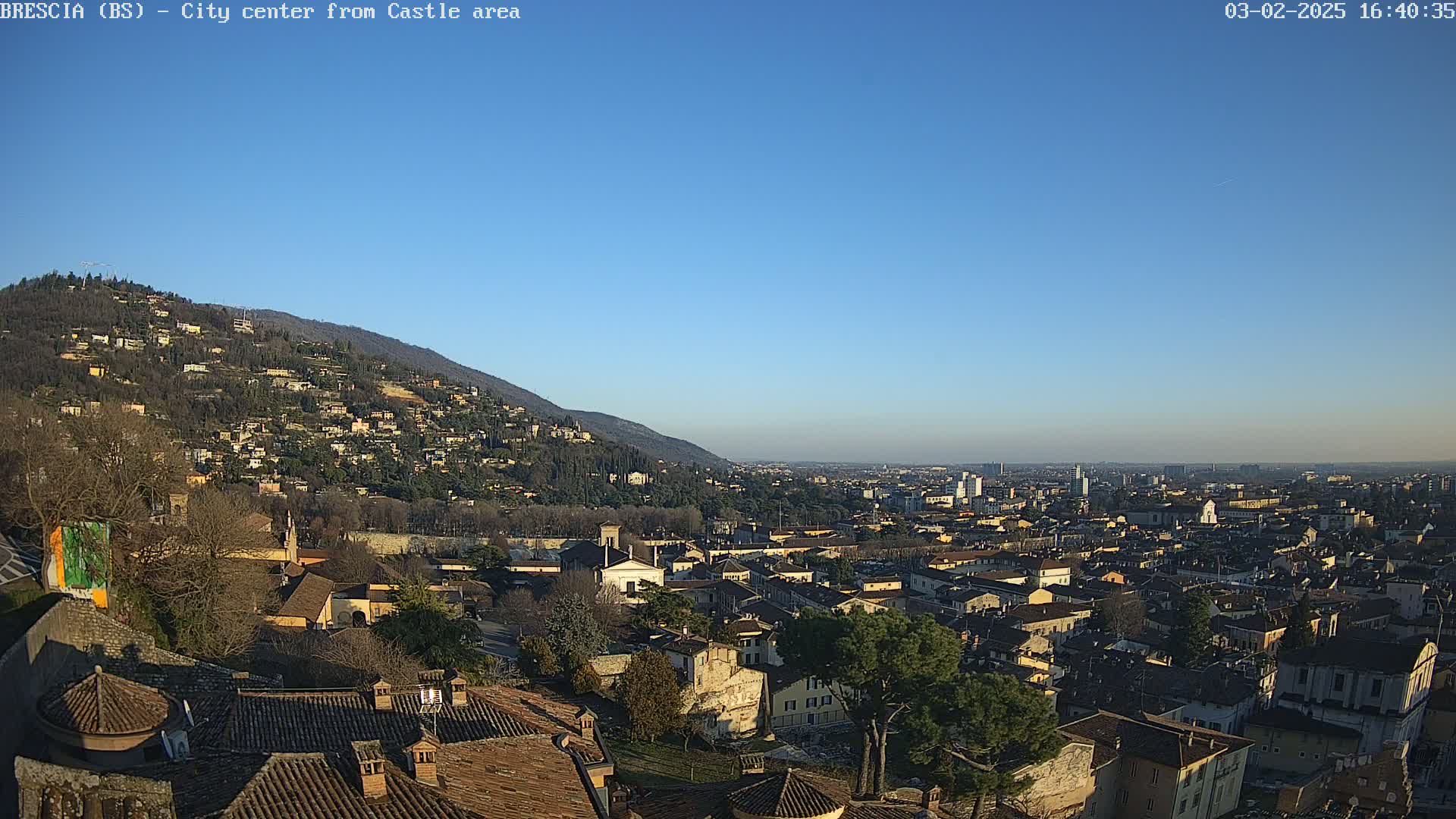 A sprawling city scene is visible from a hilltop vantage point under a clear blue sky, with buildings and houses extending to the horizon.