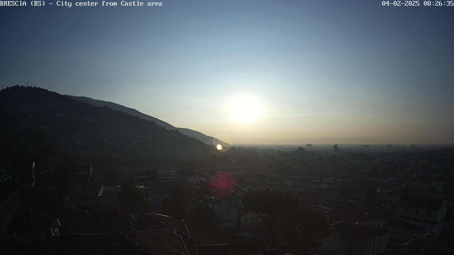 A hazy sunrise illuminates a city nestled in a valley, partially obscured by a hill in the foreground.