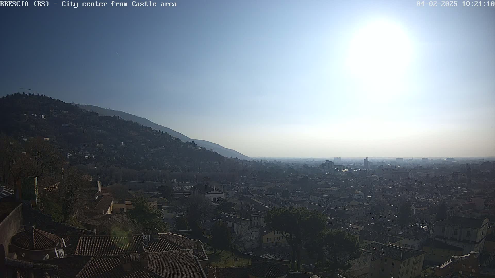 A hazy, sunny day reveals a panoramic view of a city nestled against a hillside, with many low buildings and rooftops visible from an elevated perspective.
