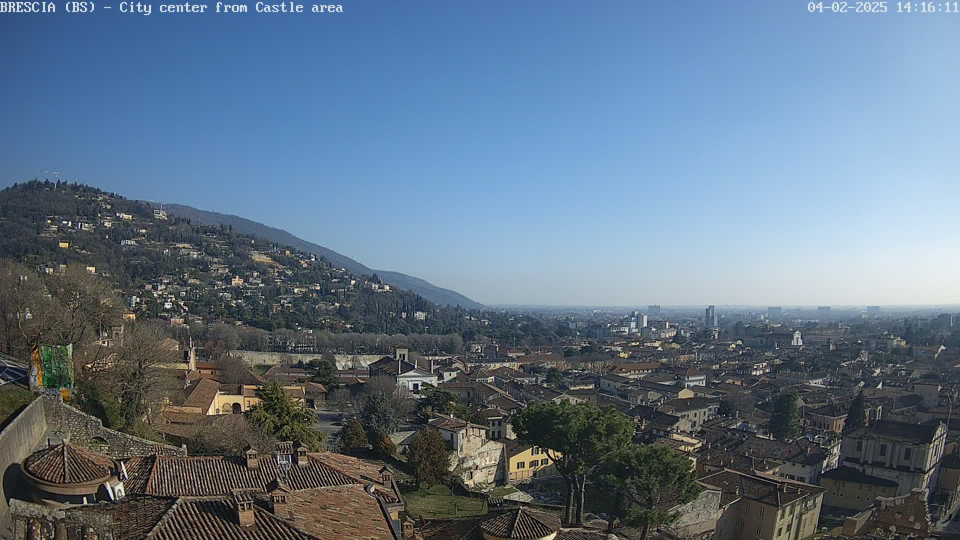 A sprawling city scene on a clear, sunny day, viewed from an elevated position overlooking numerous buildings extending to the horizon, with a hillside visible in the background.