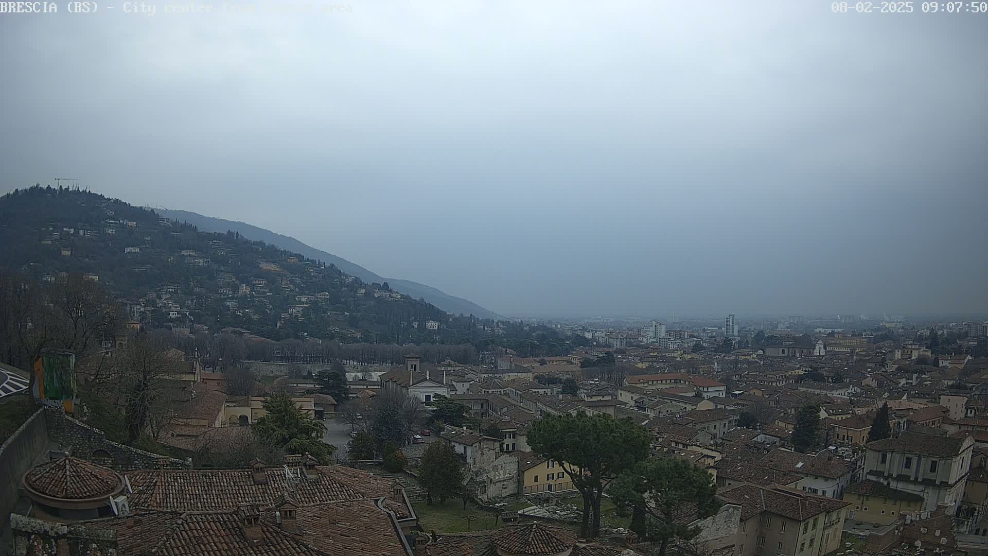 A hazy, overcast day reveals a sprawling city nestled at the foot of a hill, with numerous brown-roofed buildings extending to the horizon.