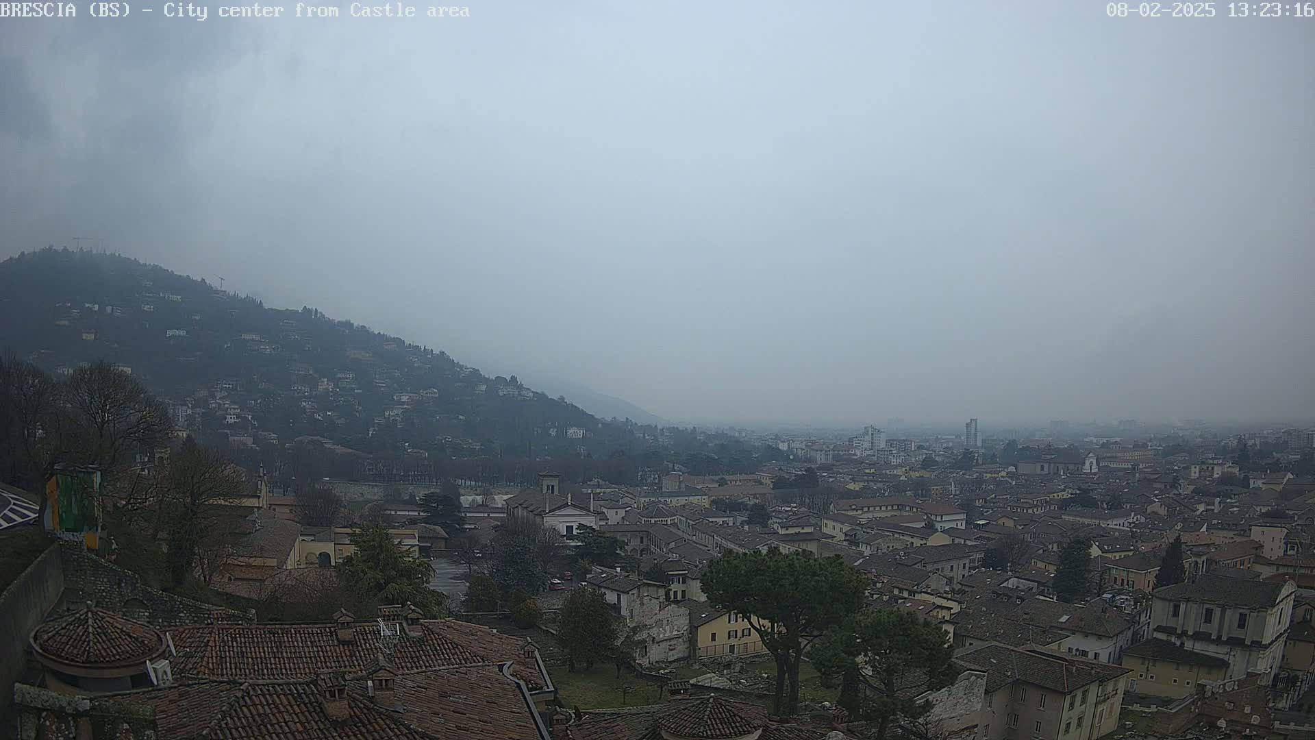 A hazy, overcast view shows a sprawling city nestled at the foot of a hill, with numerous buildings and rooftops visible from a high vantage point.