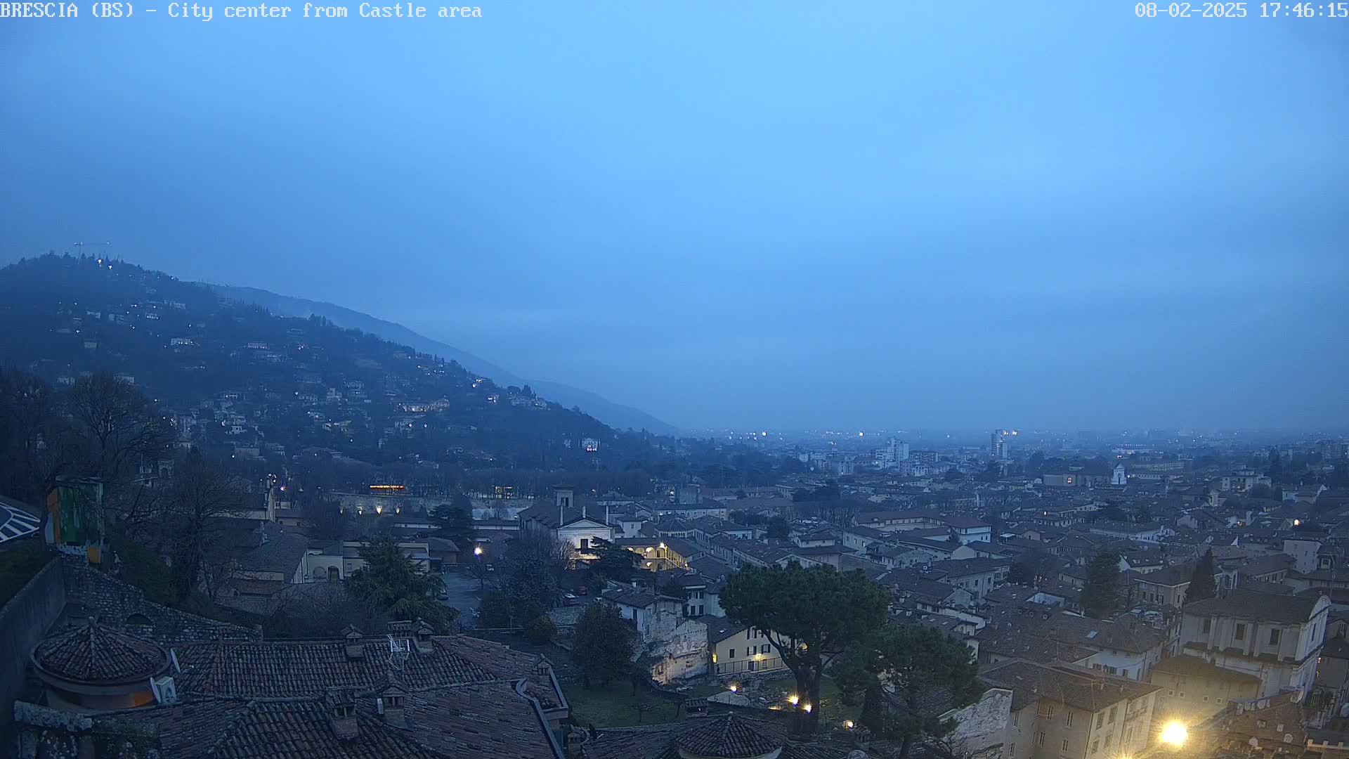 A city nestled in a valley at twilight, under a dusky blue sky, is viewed from a hilltop.