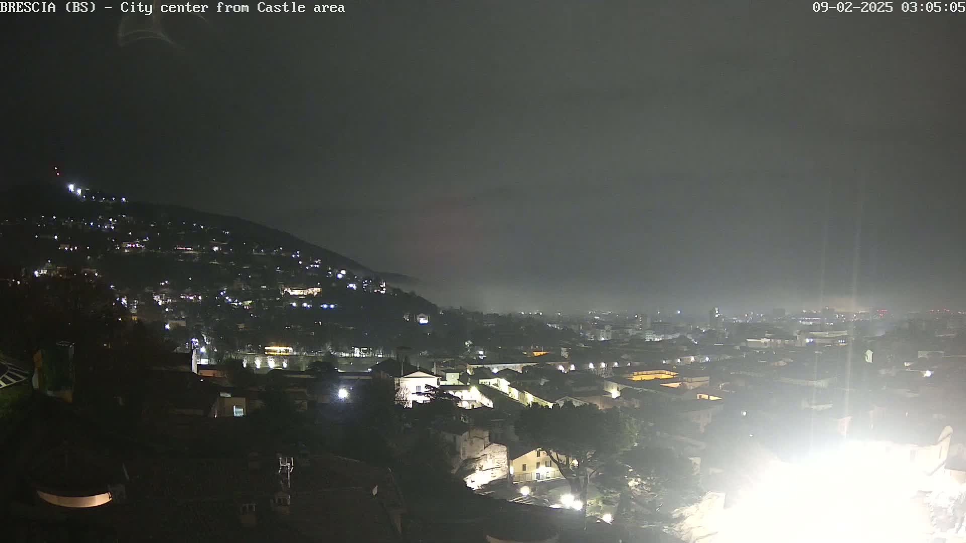 A nighttime view of a city nestled in a valley, partially obscured by fog under a dark sky.