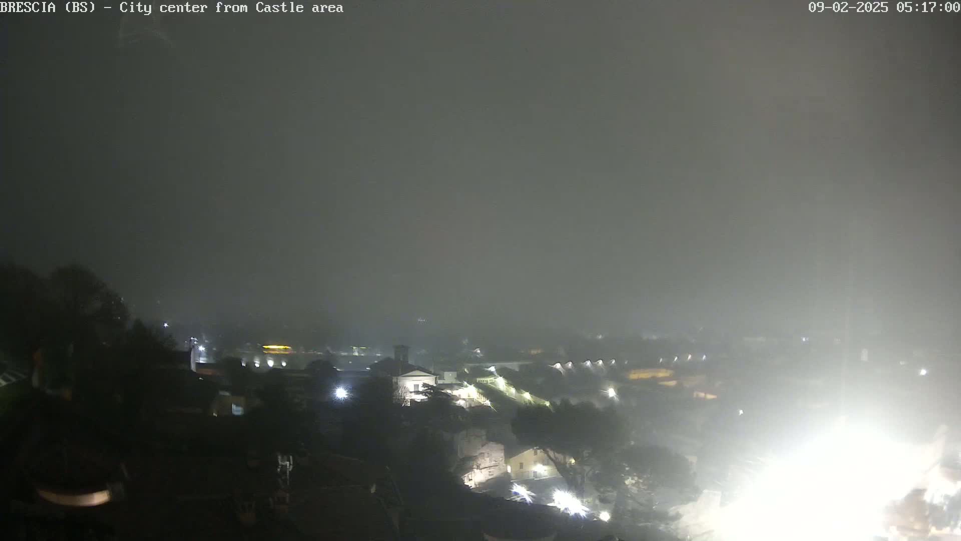A nighttime view of a city shrouded in heavy rain or fog, with dimly lit buildings and structures visible in the distance.