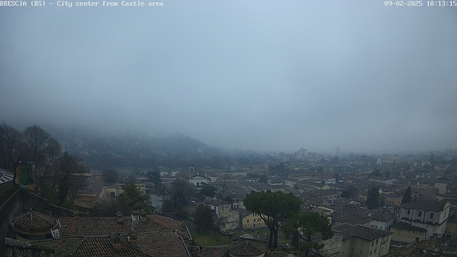 A foggy view of a city sprawling across a hill, seen from a higher vantage point with many brown-tiled rooftops visible in the foreground.