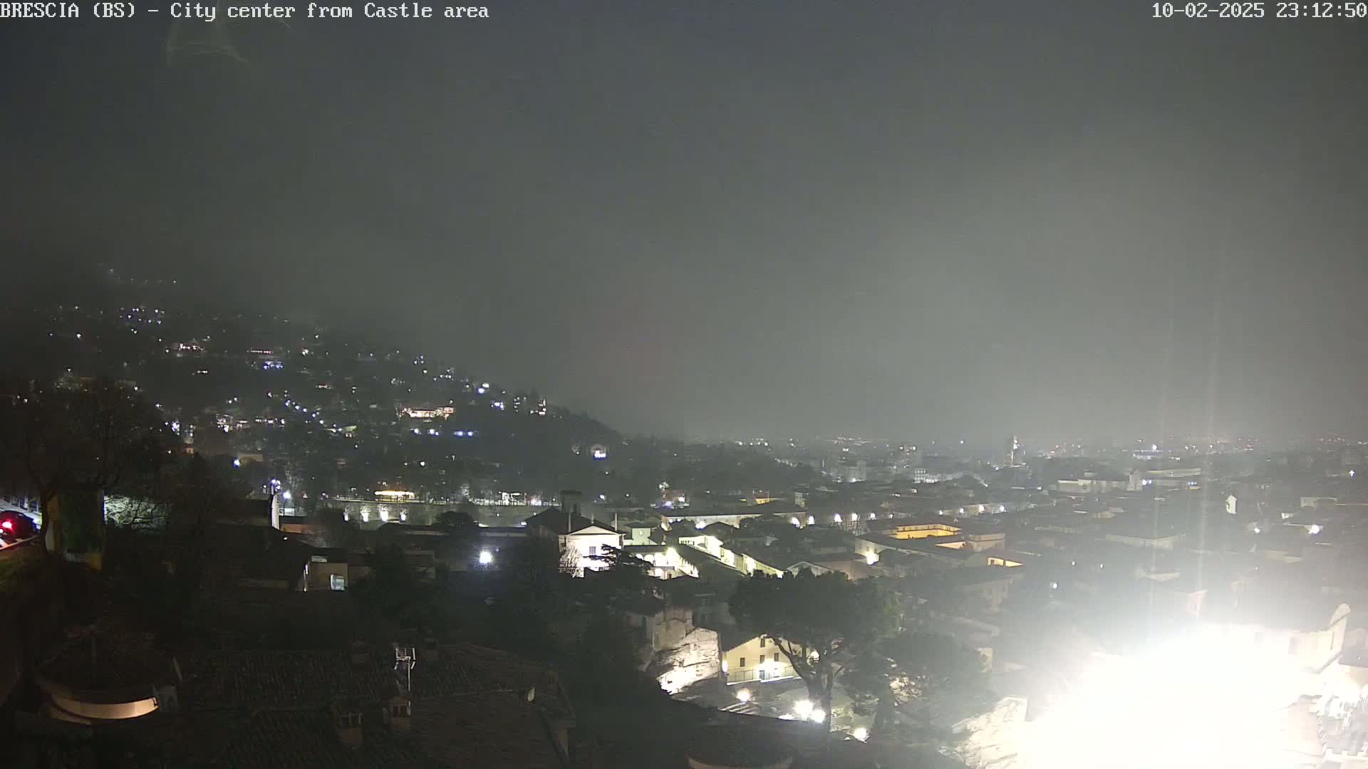 A nighttime view of a city, shrouded in fog or mist, shows a hillside with scattered lights and a lower area with densely packed buildings, also illuminated.