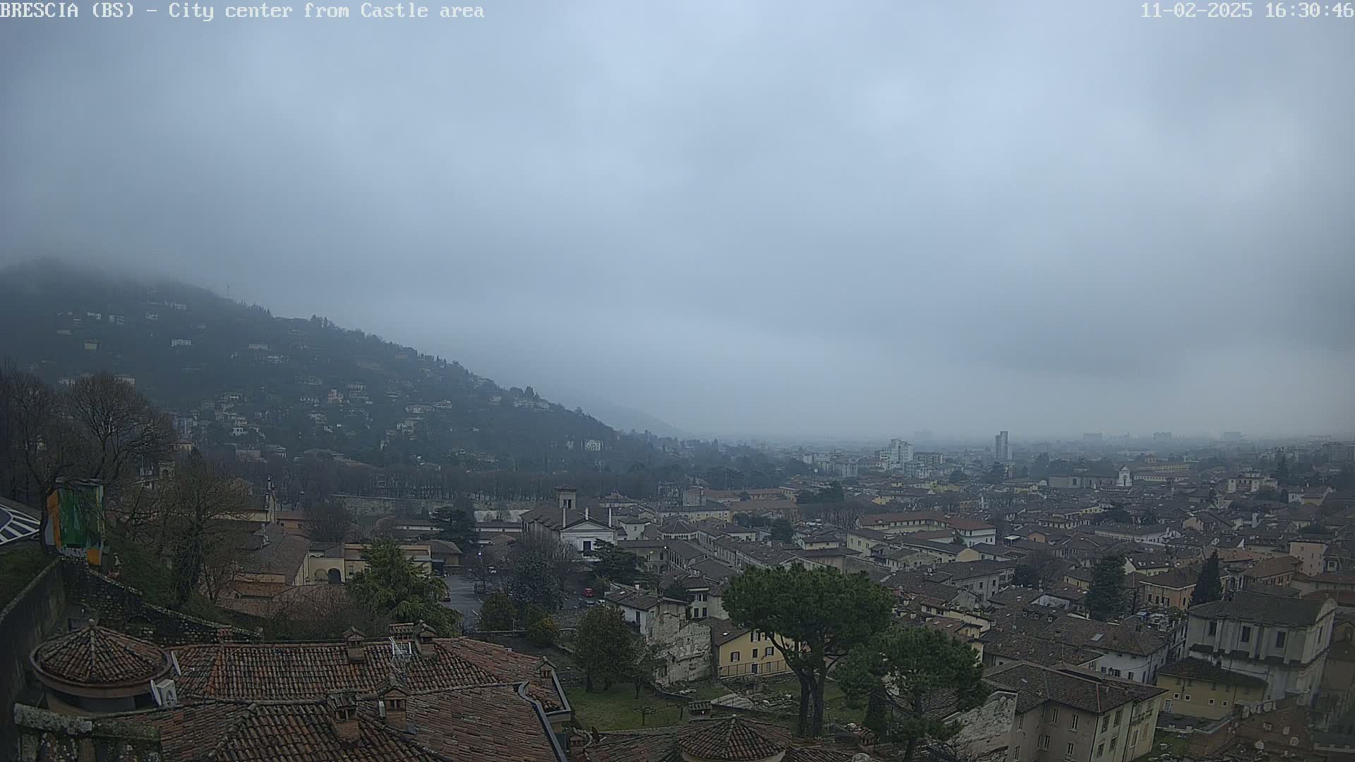 A hazy, overcast view shows a sprawling city nestled in a valley between hills, seen from a high vantage point.