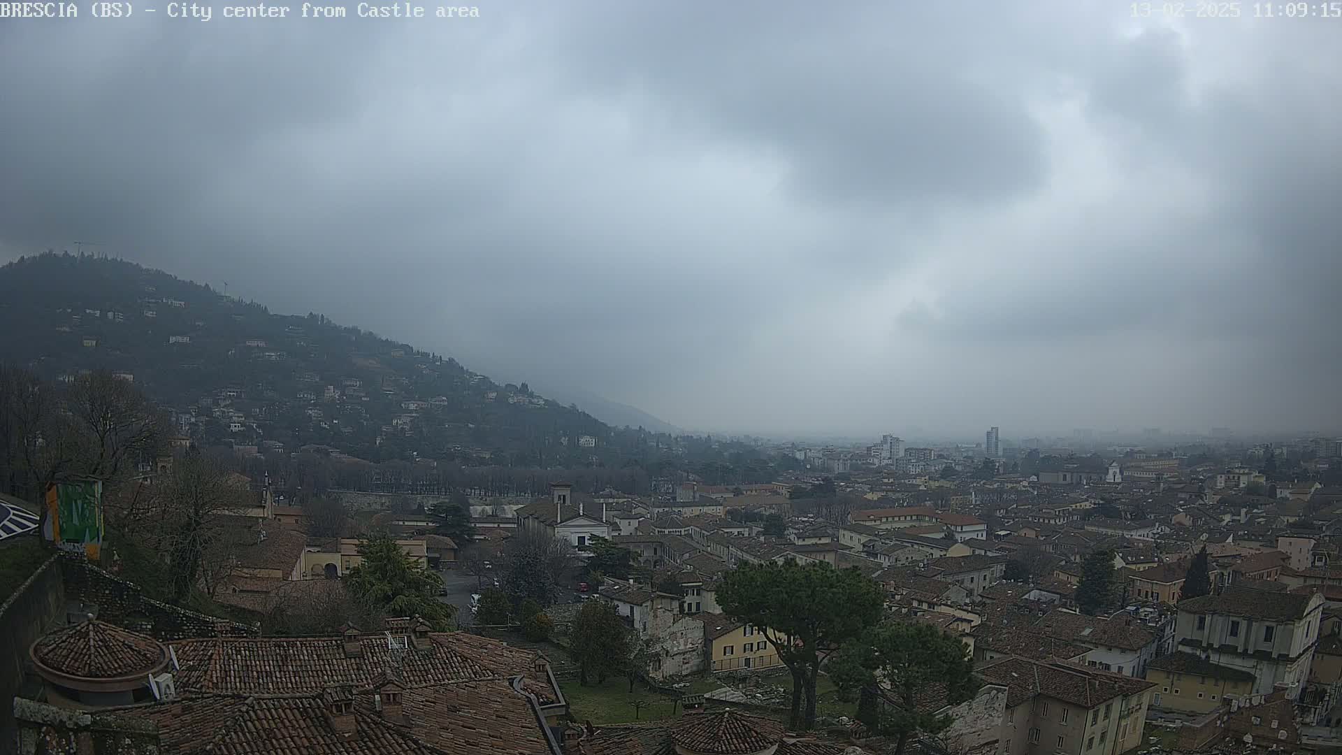 A sprawling city with many low buildings and terracotta roofs is seen from a hilltop vantage point under an overcast, hazy sky.