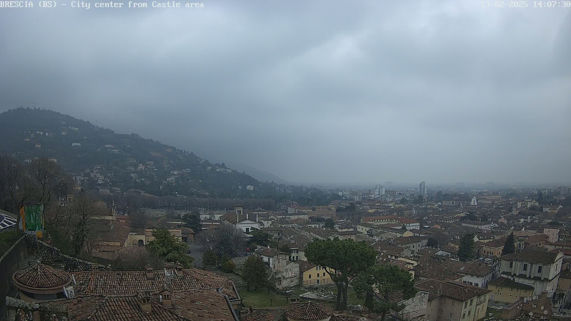 A sprawling city with red-tiled roofs nestled in a valley below a hill, all under a hazy, overcast sky.