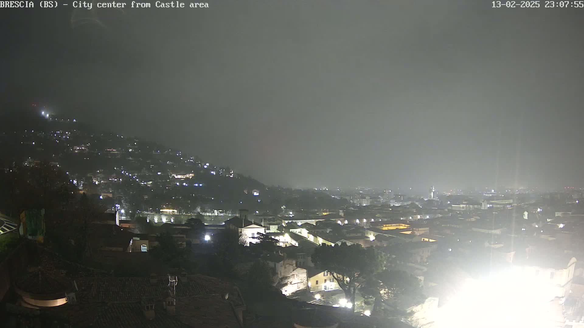 A nighttime view of a city nestled into a hillside under hazy or foggy conditions, with the lights of buildings visible across the landscape.