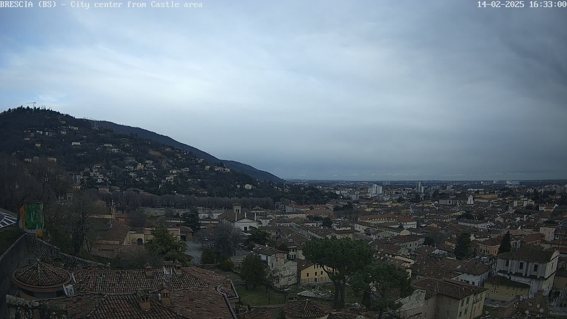 A view from a hilltop shows a city of brown-roofed buildings under a cloudy sky.