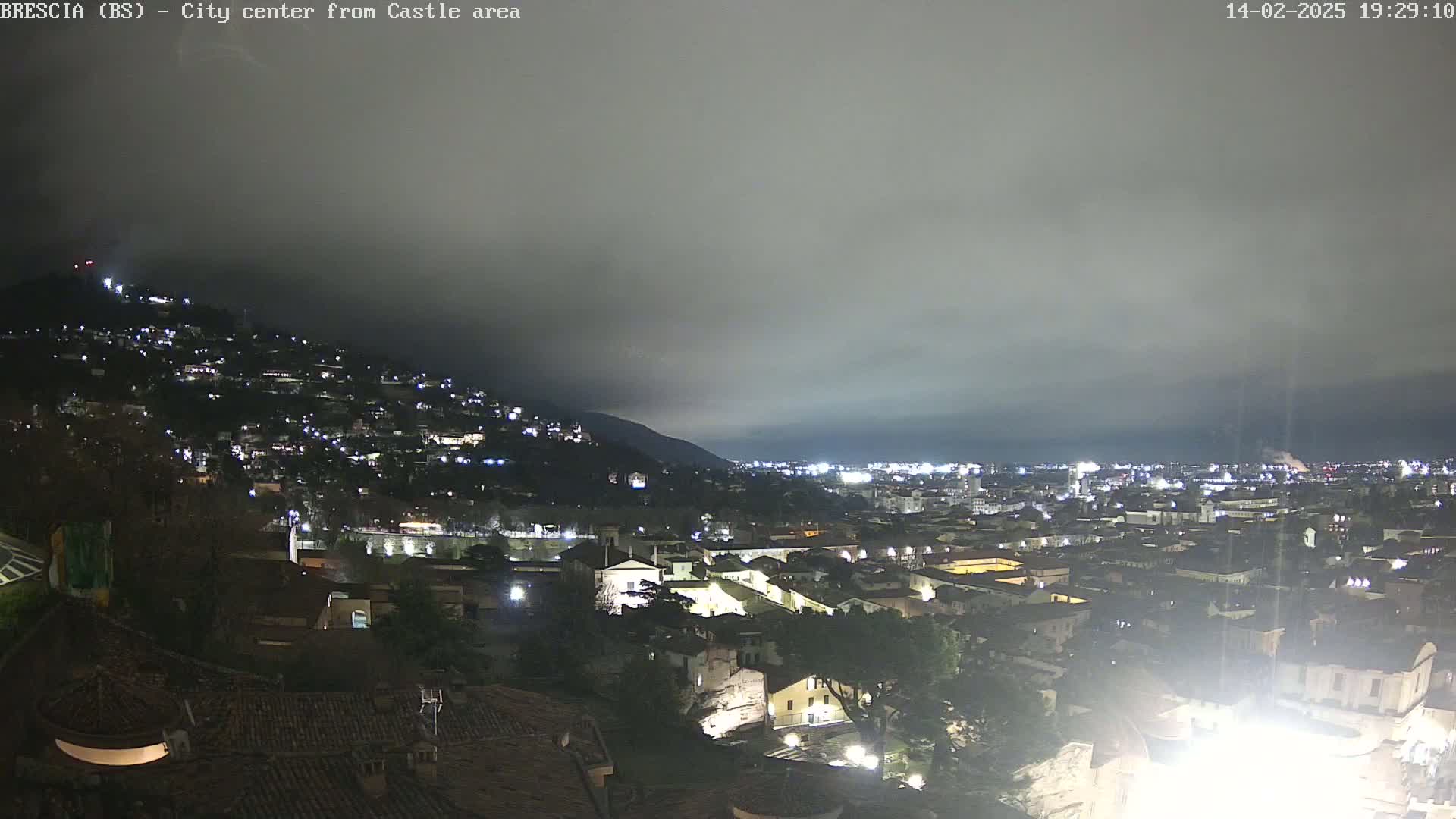 A nighttime view of a city nestled in a valley under a cloudy sky, with lights illuminating buildings and streets.