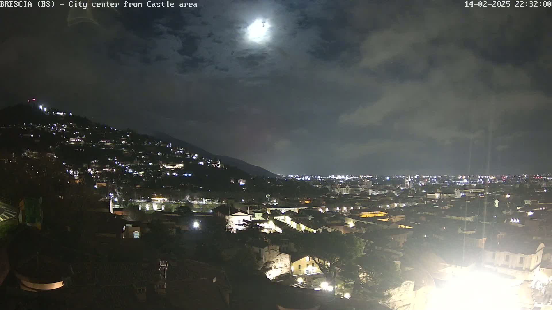 A nighttime view of a city nestled in a valley under a partly cloudy sky and a bright moon.