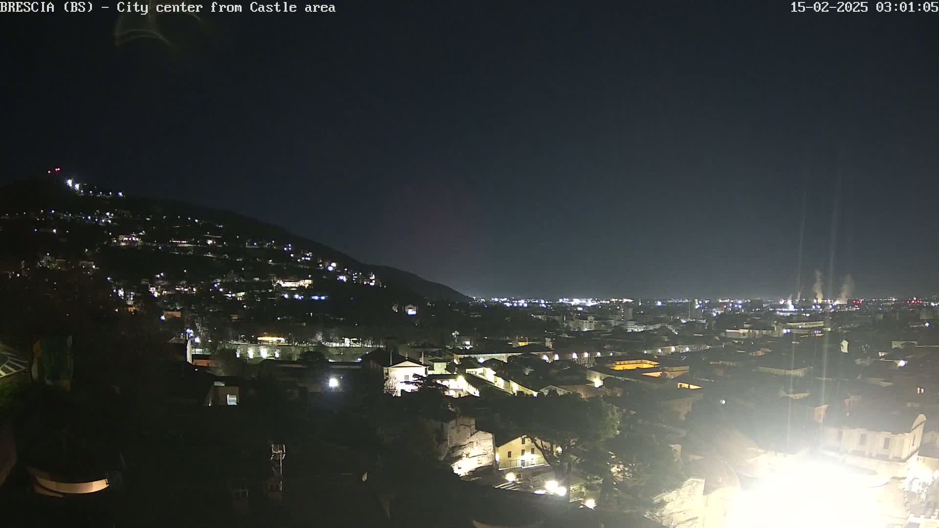 A nighttime, long shot of a city nestled against a hill, with the lights of buildings illuminating the landscape under a dark, clear sky.
