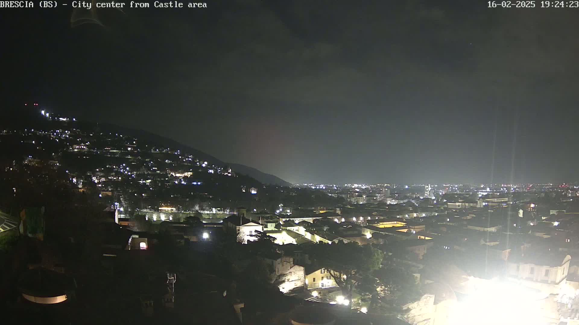 A nighttime view of a city nestled against a hill, under a mostly cloudy sky.