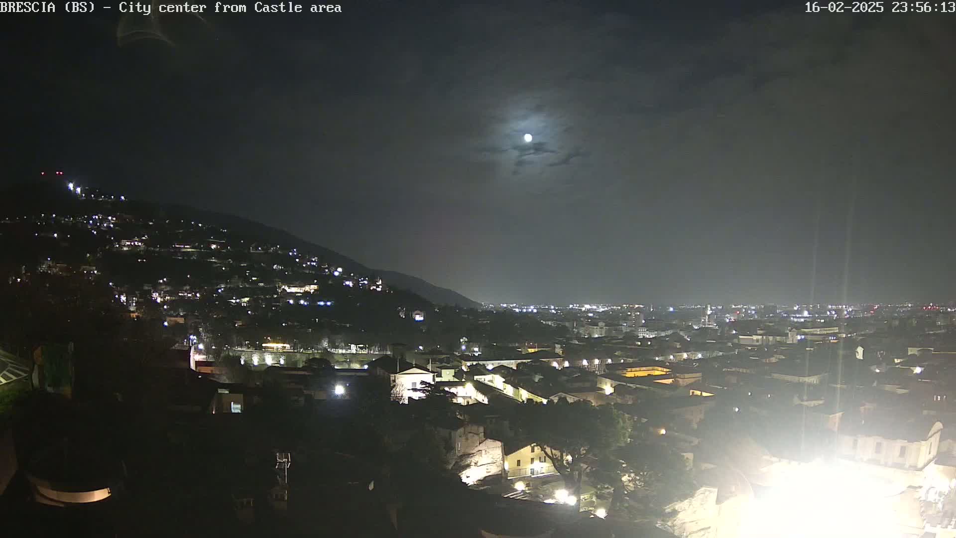A nighttime, mostly cloudy view of a city nestled against a hill, illuminated by many streetlights and a bright moon.