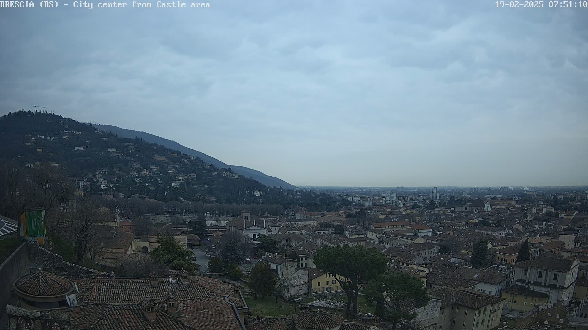 A sprawling city with red-tiled roofs nestled beneath a hill, viewed on an overcast day.